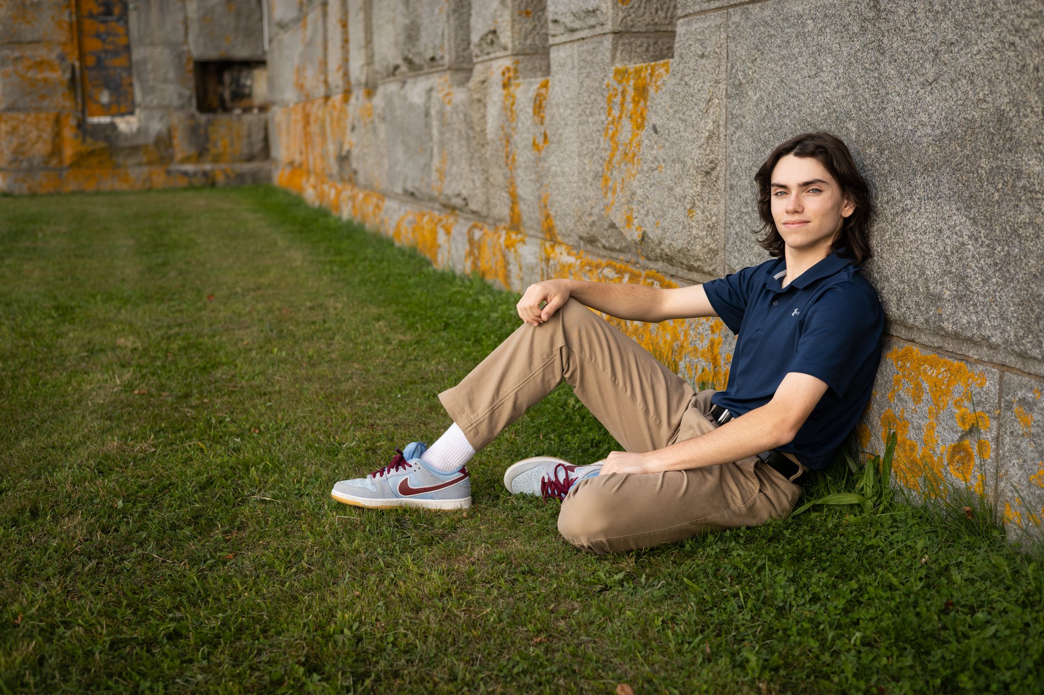 A young man with shoulder-length dark hair sitting on grass against a large stone wall with patches of yellow lichen, wearing a navy blue polo shirt, khaki pants, and white sneakers with maroon accents.