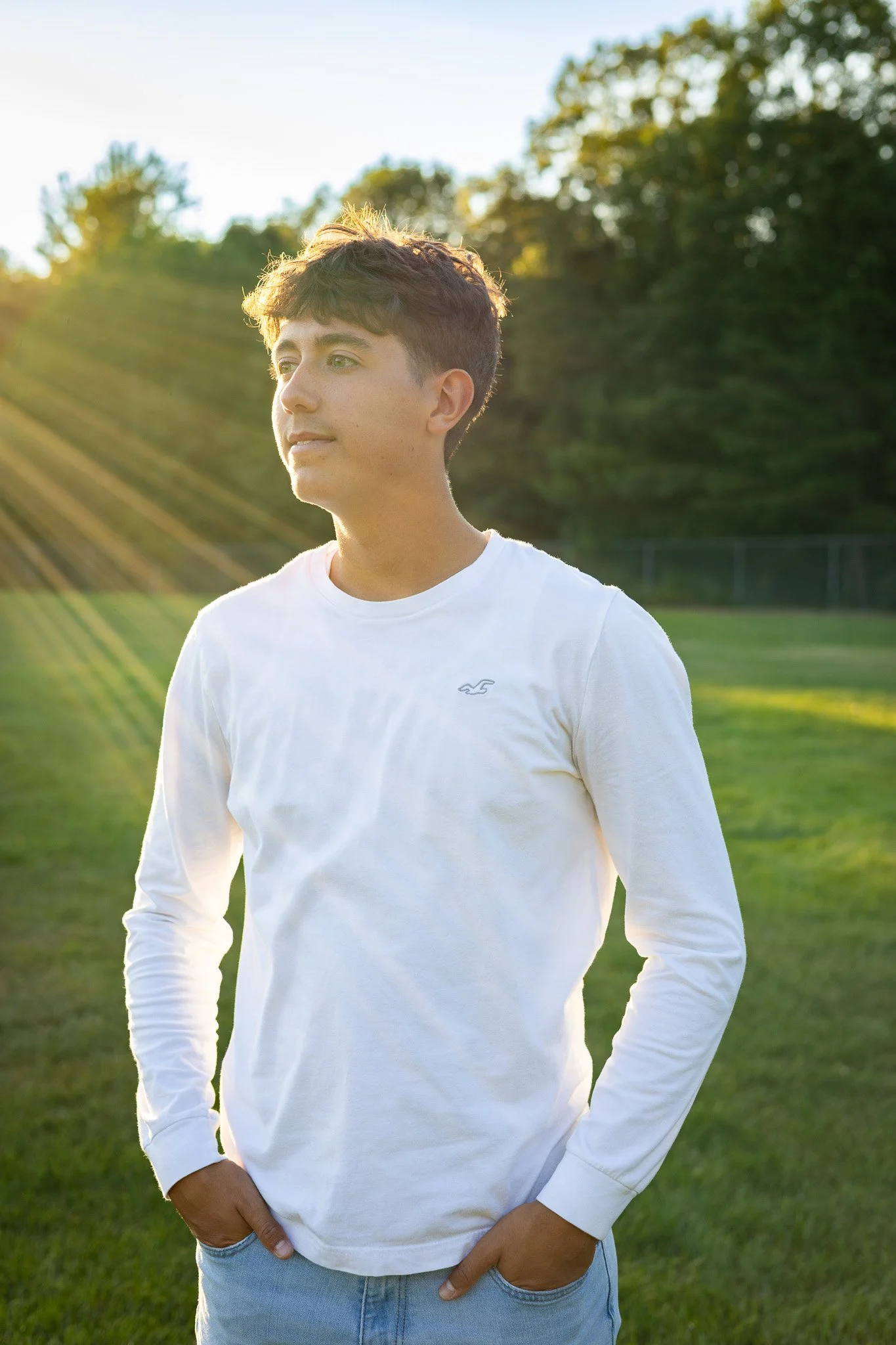 Young man standing outdoors on grass field during sunset, wearing a white long-sleeve shirt and jeans, with trees in the background.
