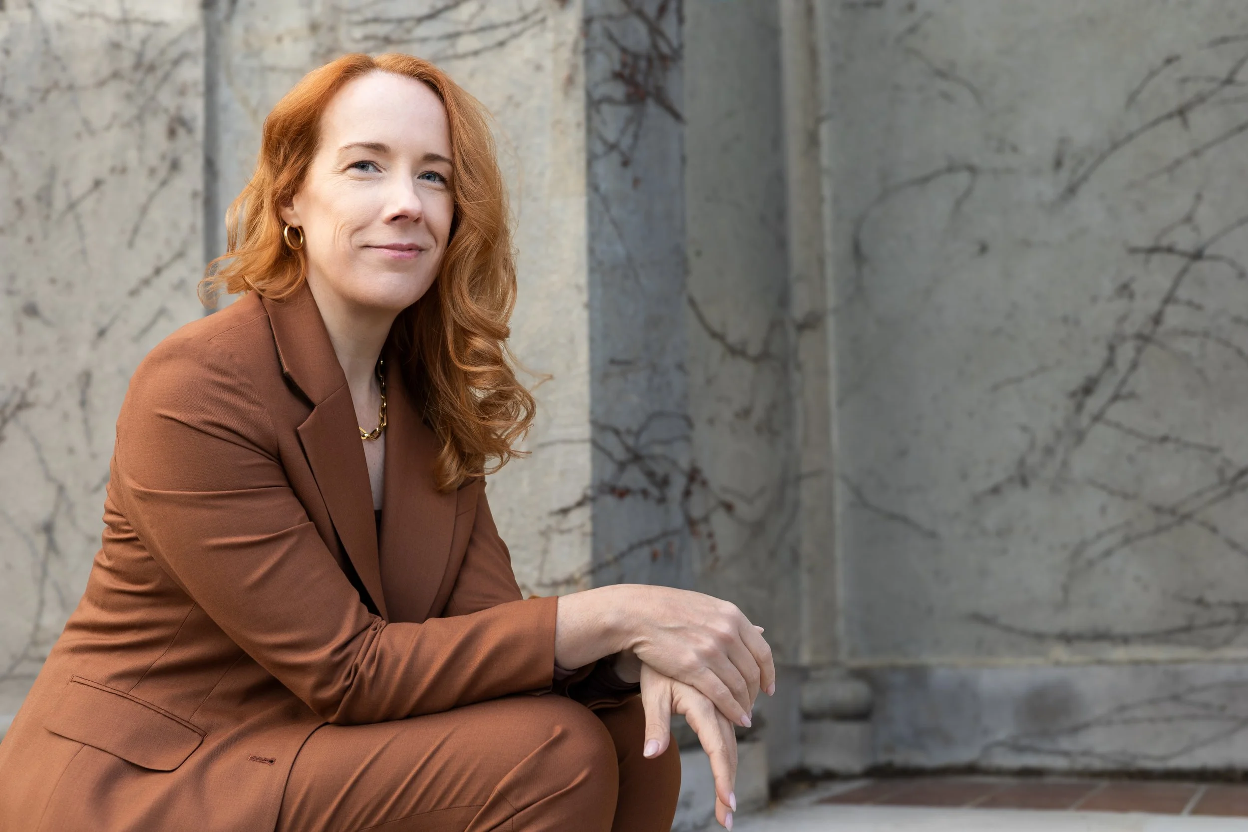 A woman with red hair and gold hoop earrings, wearing a brown blazer, sitting outdoors in front of a stone wall with vines.