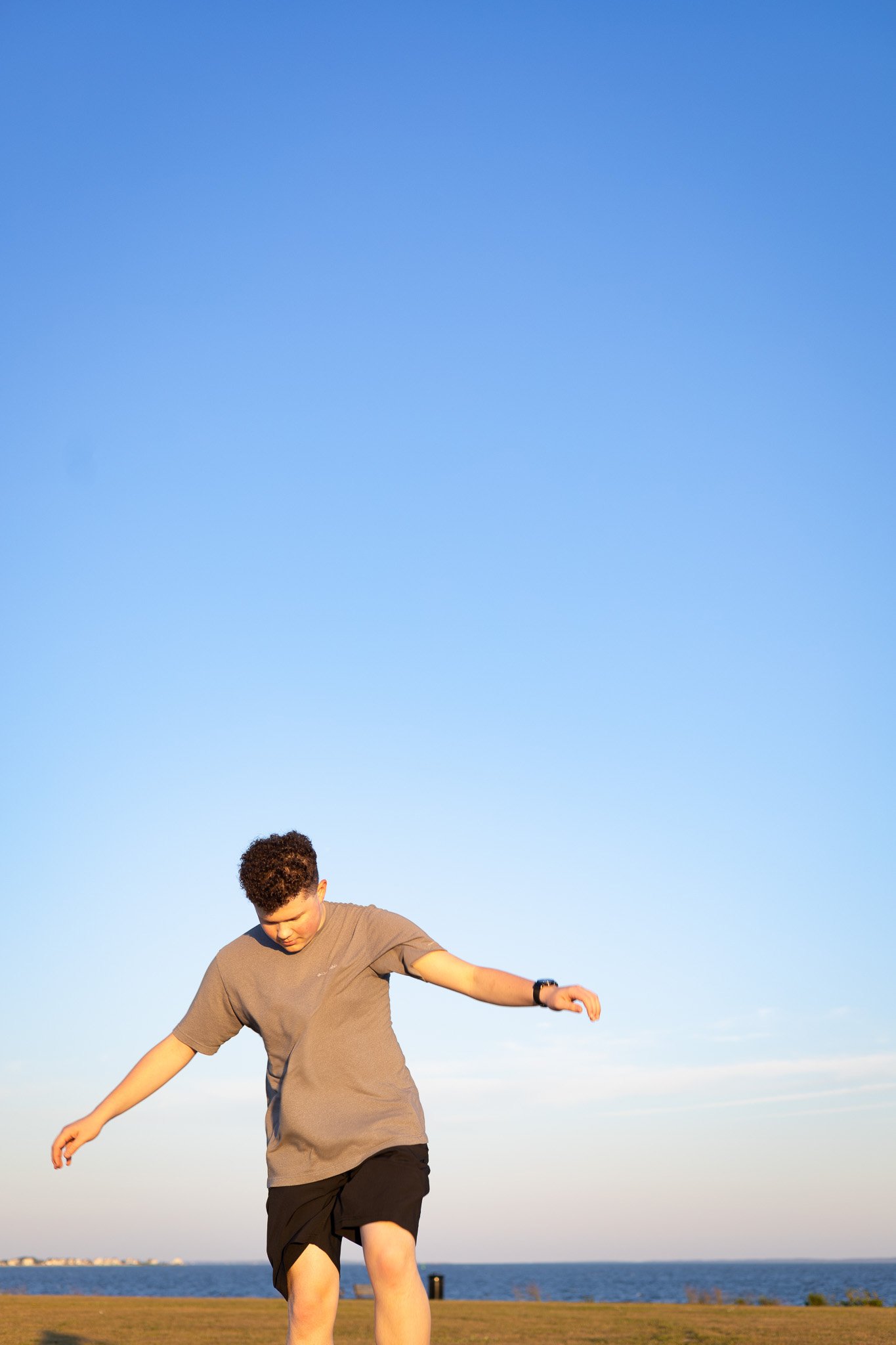 A young man with curly hair walking on grass near a body of water, under a clear blue sky.