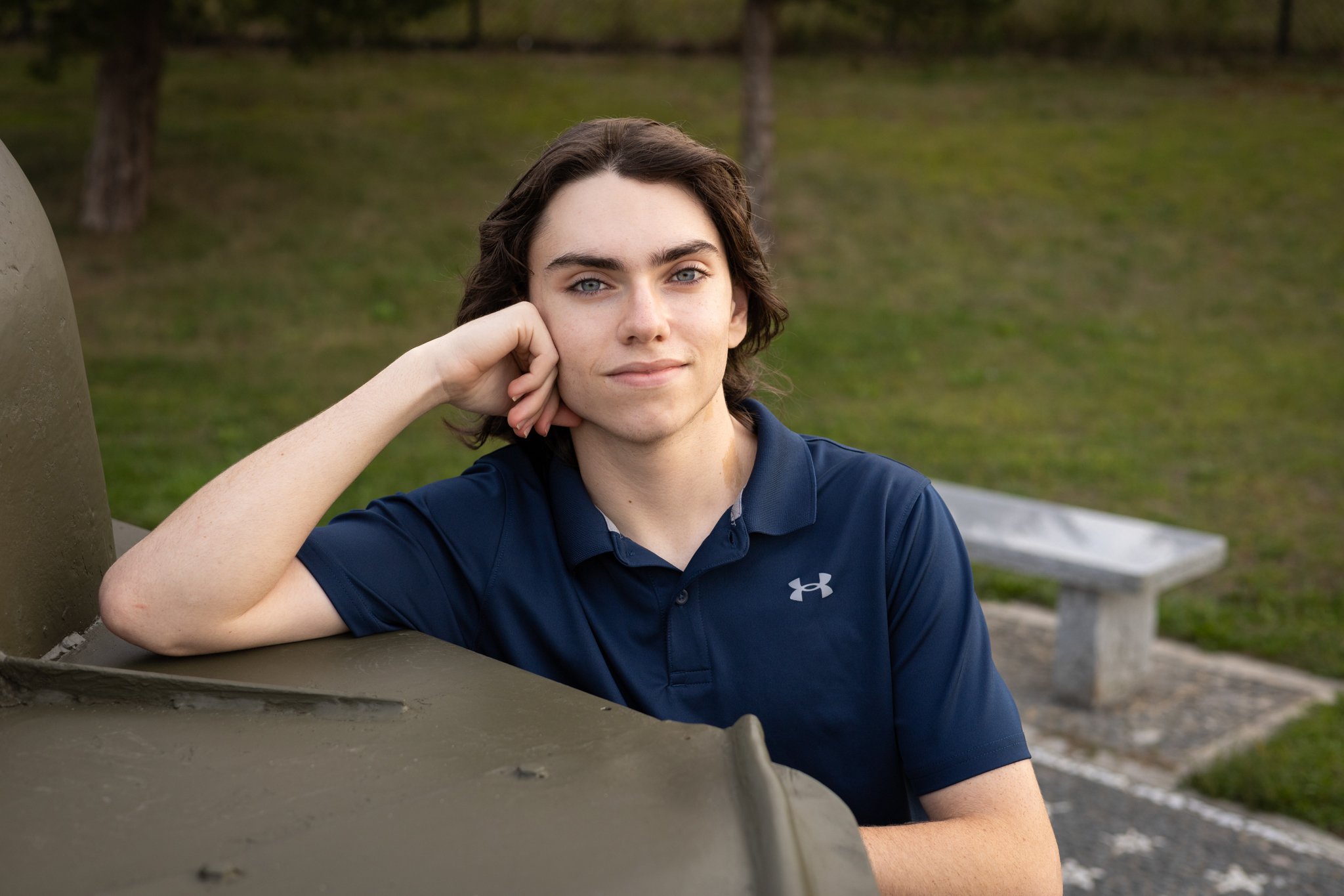 A young woman with shoulder-length dark hair and blue eyes, wearing a navy blue Under Armour polo shirt, sitting on a park bench with her head resting on her hand, outdoors with trees and grass in the background.