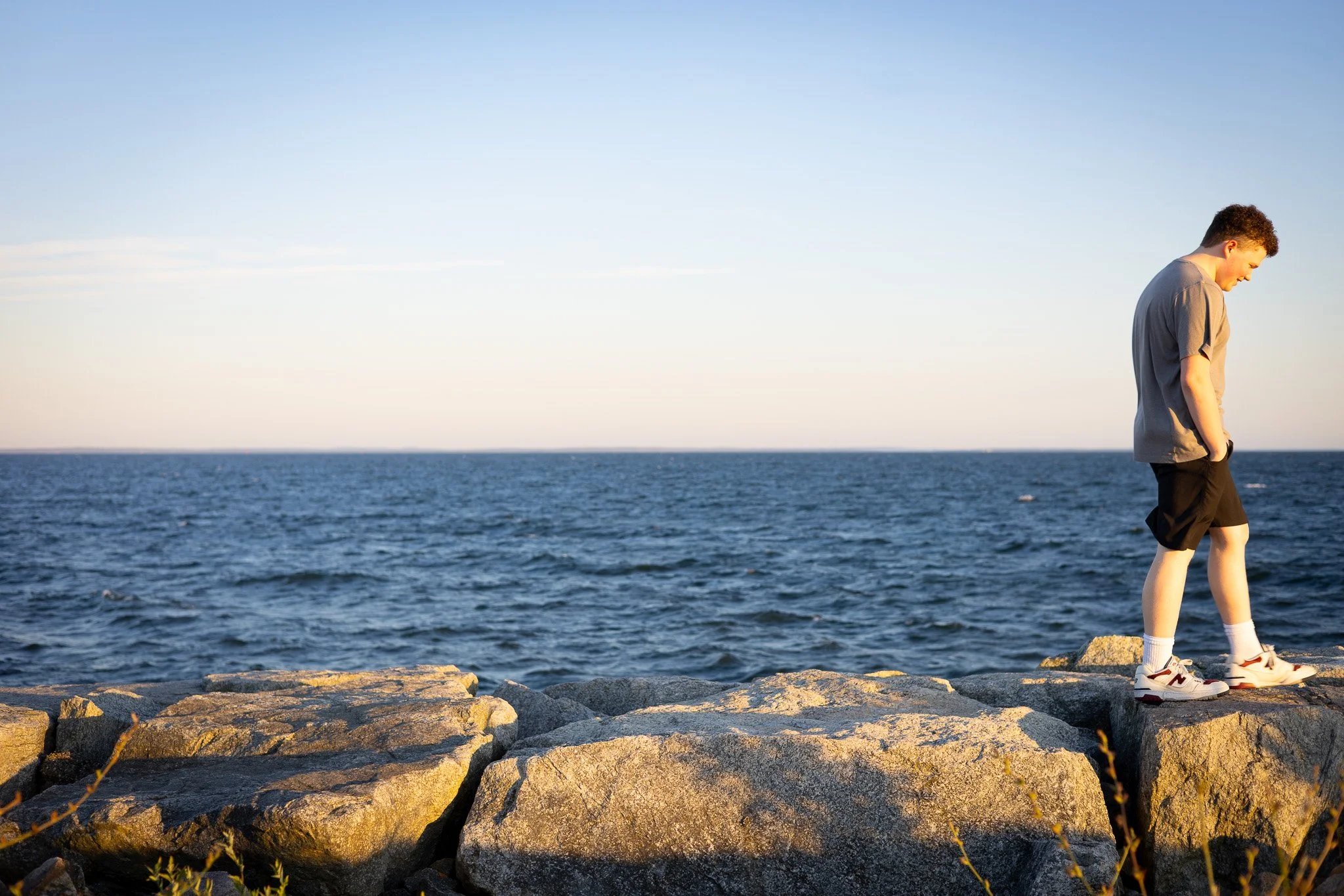 A young male with curly hair wearing a gray t-shirt, black shorts, and sneakers stands on a rocky shoreline, looking down at the ground with their hands in their pockets, by the sea during sunset.