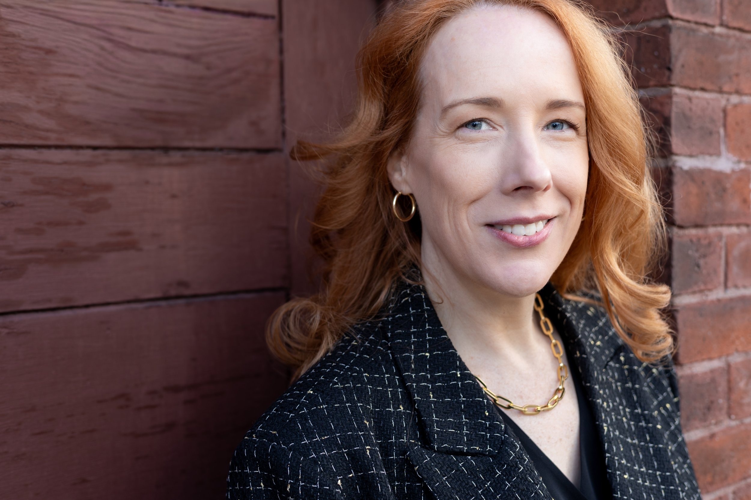 A woman with red hair and blue eyes smiling, wearing gold hoop earrings, a black and gold patterned blazer, a black top, and a gold chain necklace, standing in front of a wooden wall and brick wall.