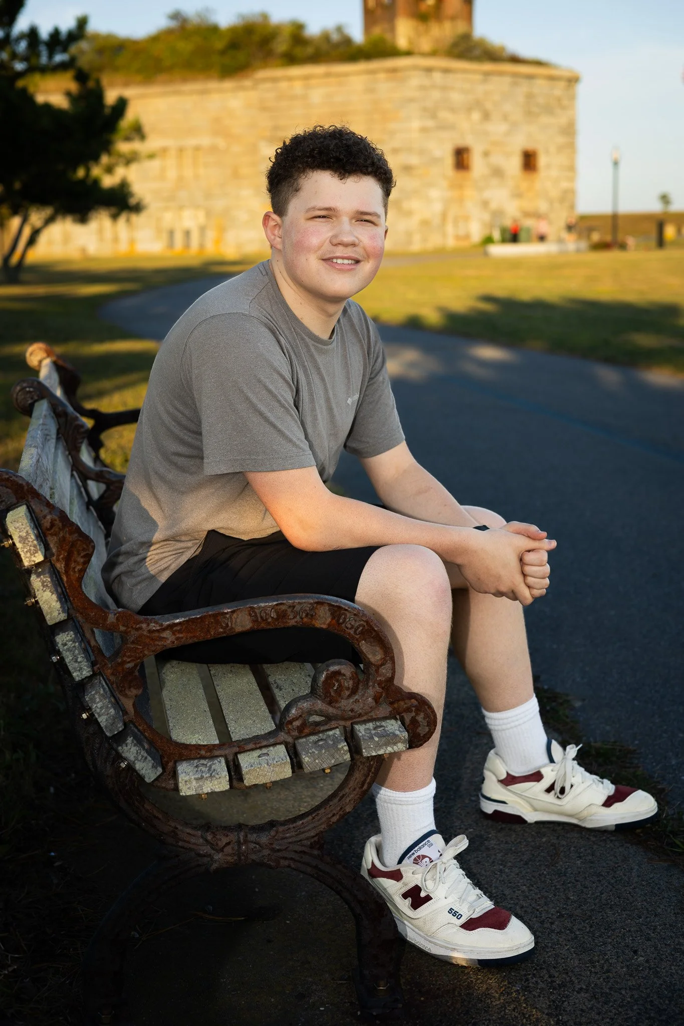 A male teen with curly hair sitting on a park bench during late afternoon or early evening. He is wearing a gray t-shirt, black shorts, white socks, and sneakers. The sunlight illuminates the scene with warm tones. The background features an historic