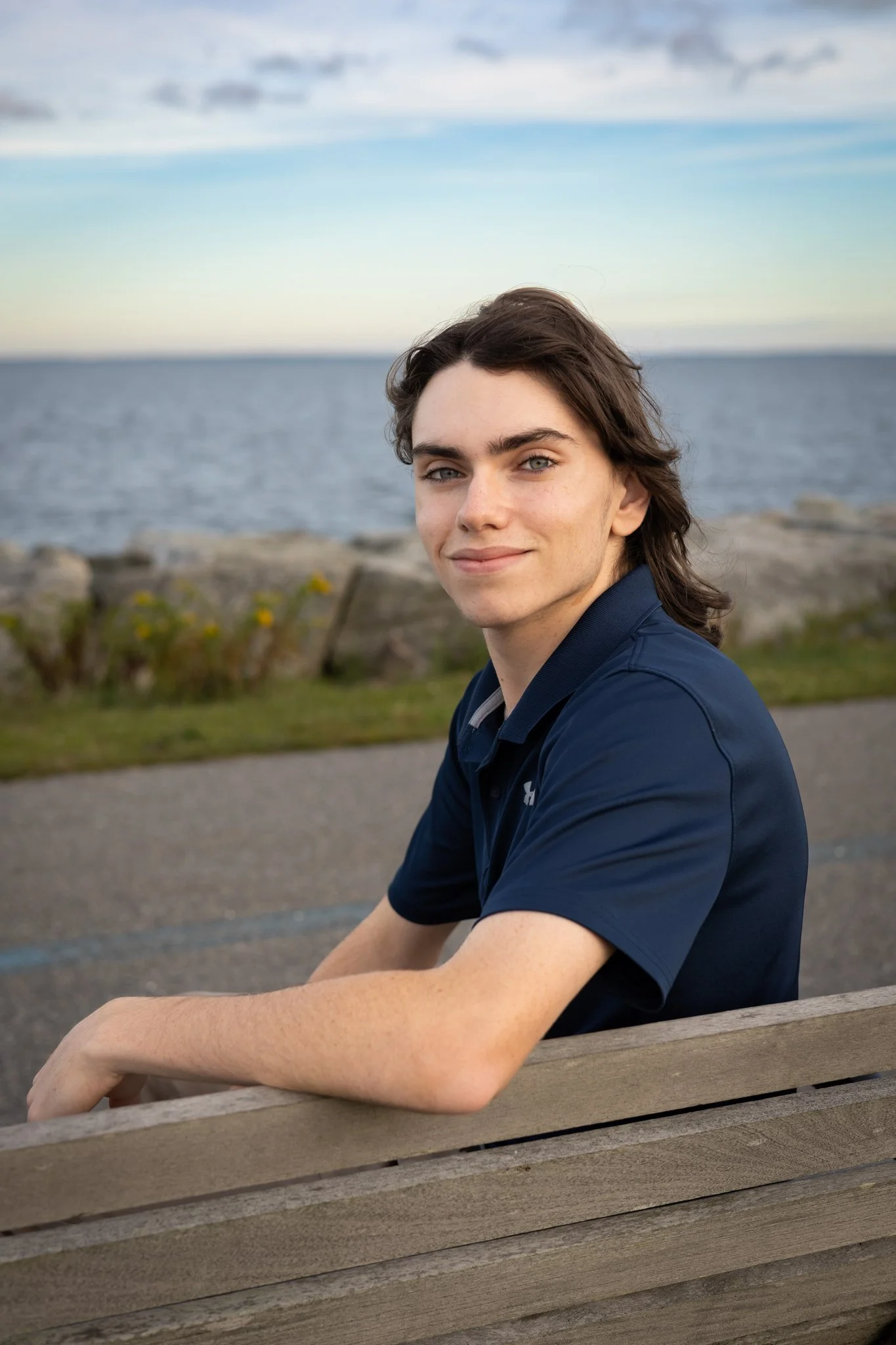 Young man with dark brown hair and blue eyes sitting on a wooden park bench near the water, smiling at the camera on a partly cloudy day.