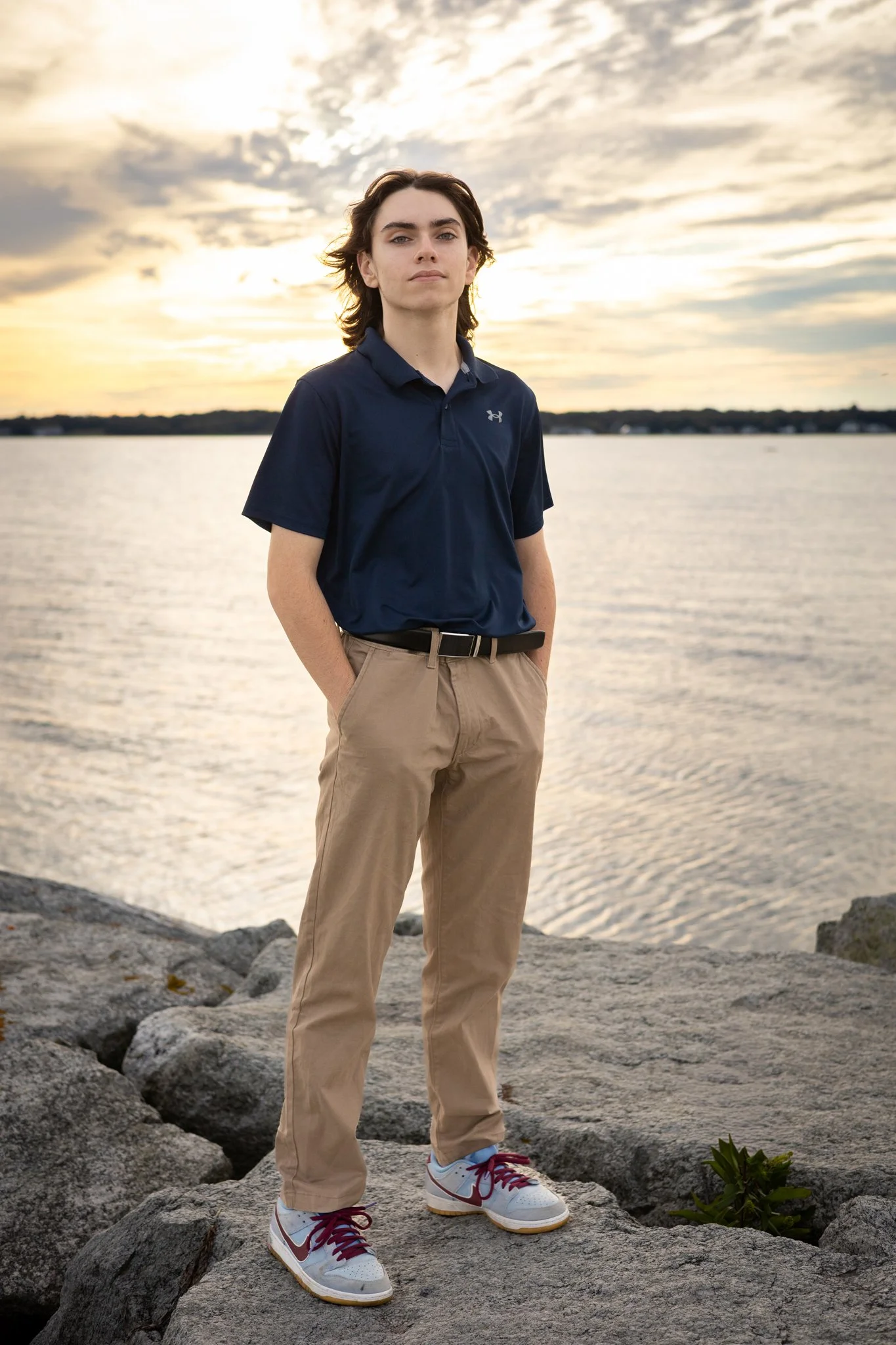 A young male standing on large rocks by a body of water during sunset, wearing a navy blue polo shirt, beige pants, and white sneakers with red laces.