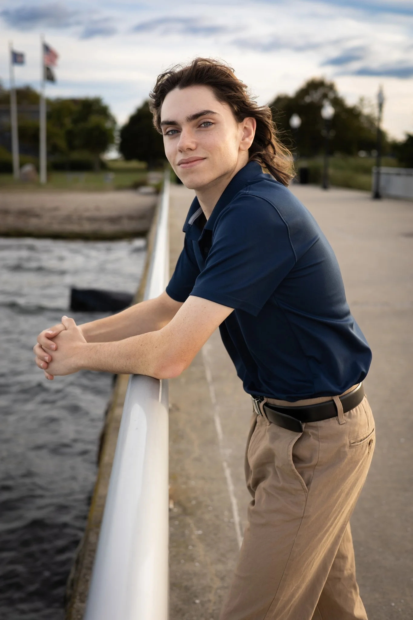 A young man with wavy brown hair and fair skin leaning on a white railing at a scenic outdoor location with water, trees, and American flags in the background.