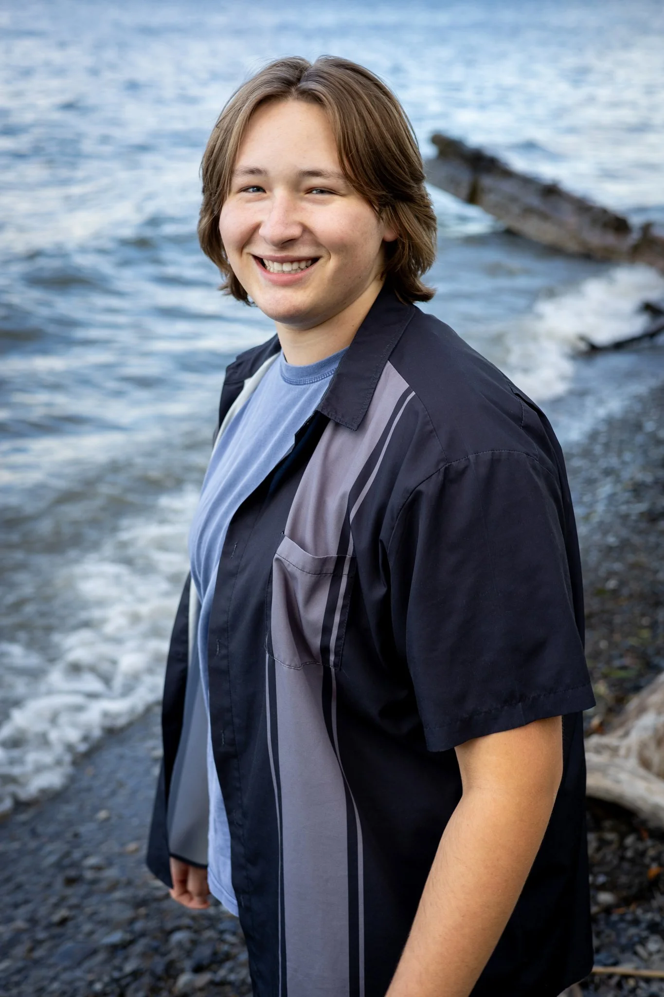 A young man with shoulder-length brown hair smiling at the camera, standing by the water at a rocky beach.