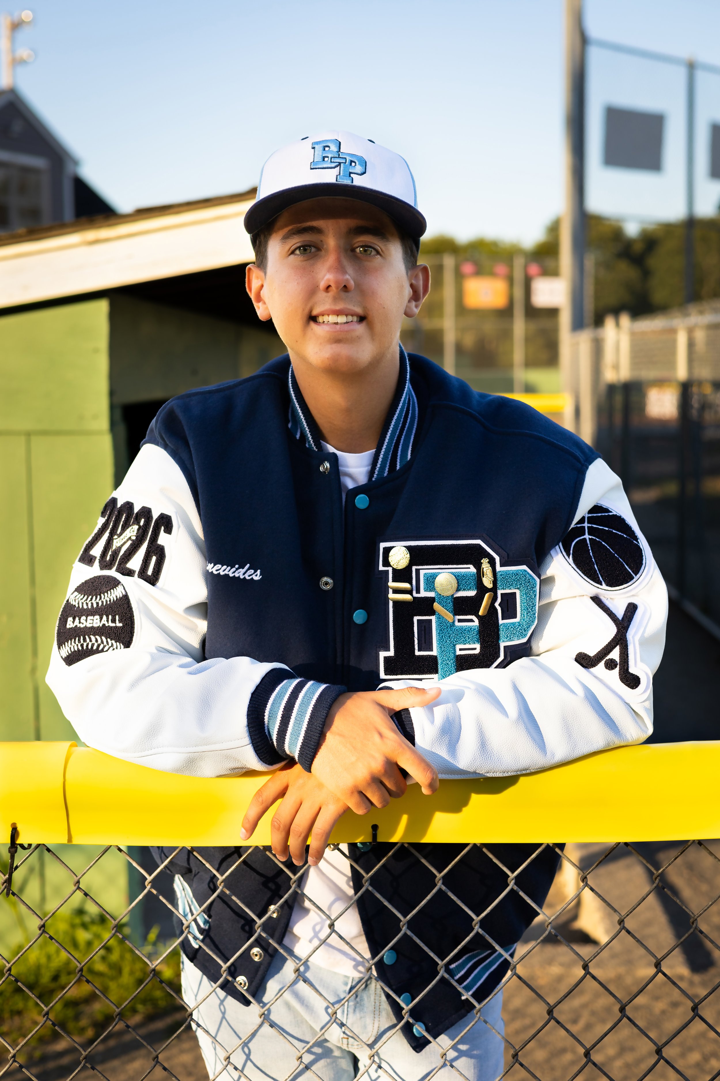 A young man in a varsity letter jacket and baseball hat leaning on a chain-link fence at a baseball field during sunset.