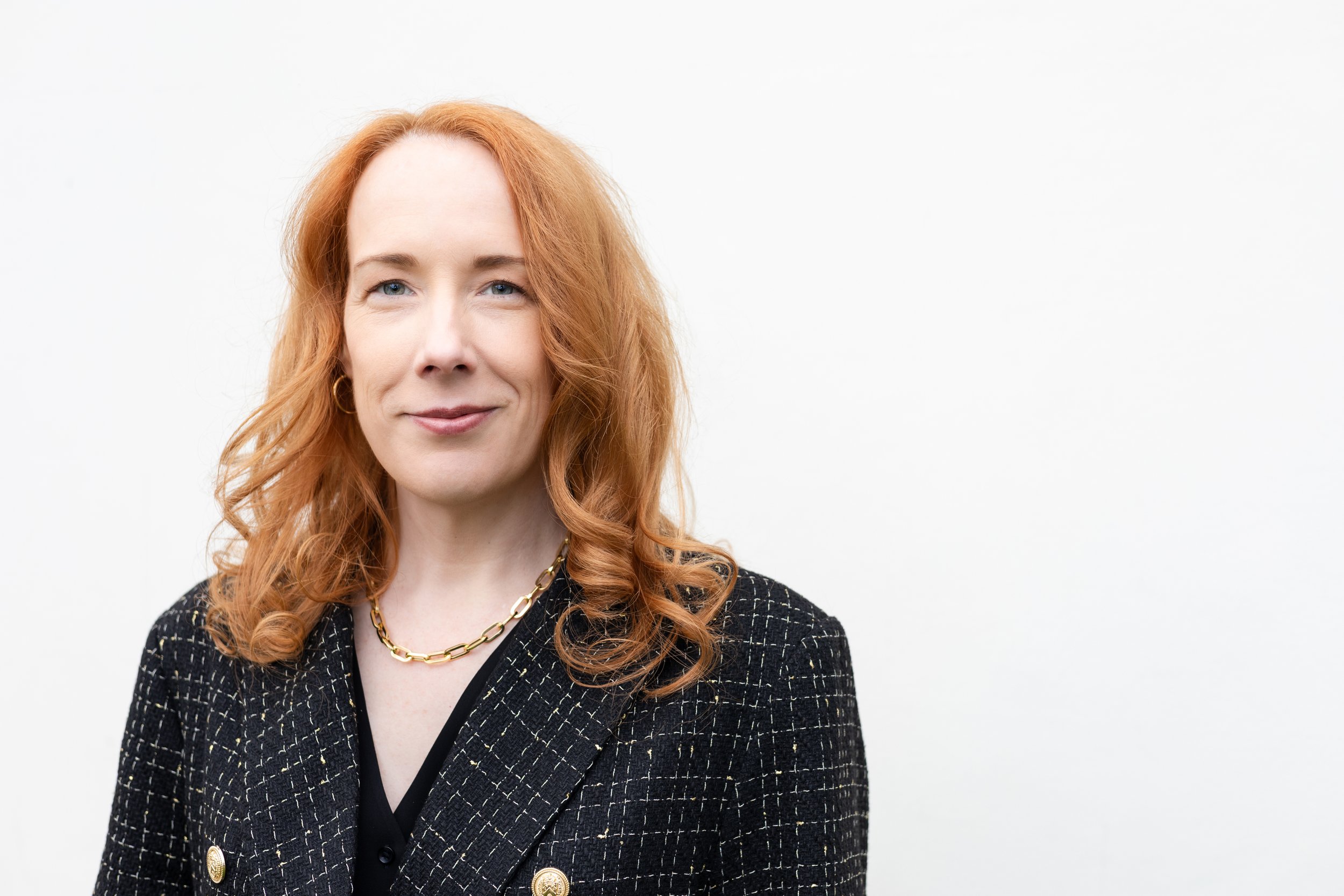 A woman with long red hair wearing a black blazer with a checkered pattern, a black top, gold hoop earrings, and a gold chain necklace standing against a plain white background.