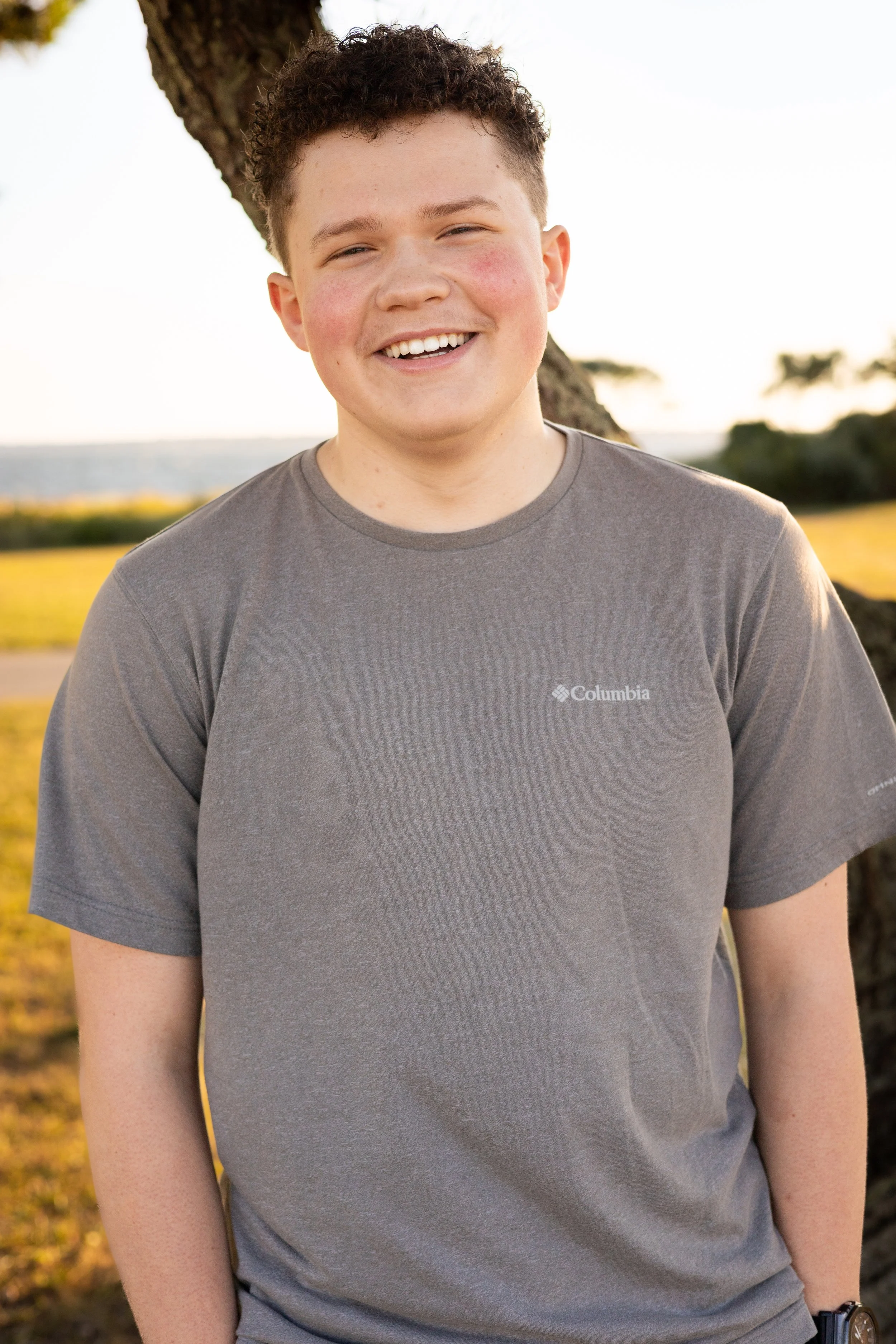 A young person with short curly hair smiling outdoors, wearing a T-shirt, standing next to a tree with a grassy area, water, and trees in the background during daytime.