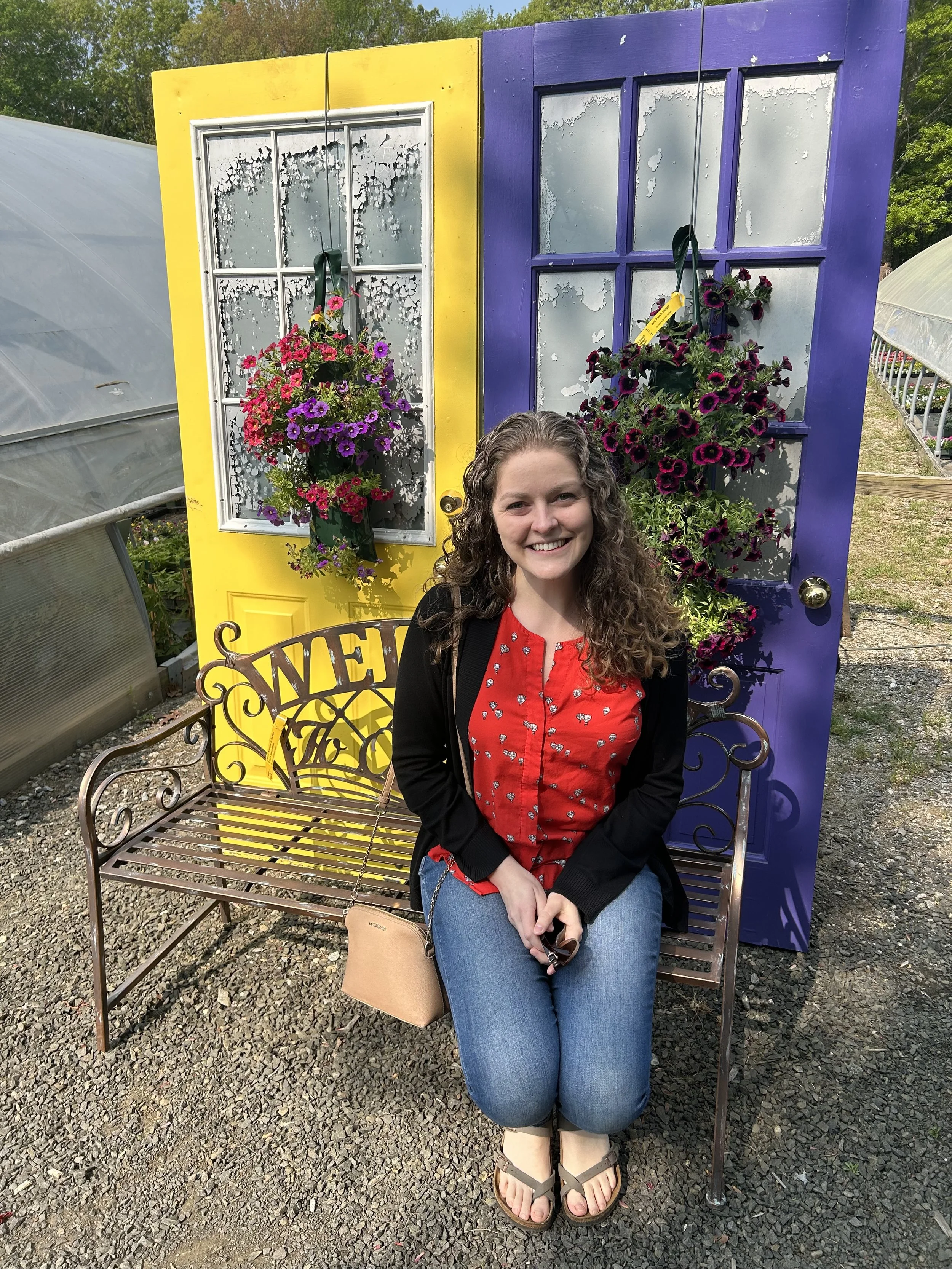 A woman with long curly hair is sitting on a bench in front of a yellow door and a purple door, each with flower baskets on them