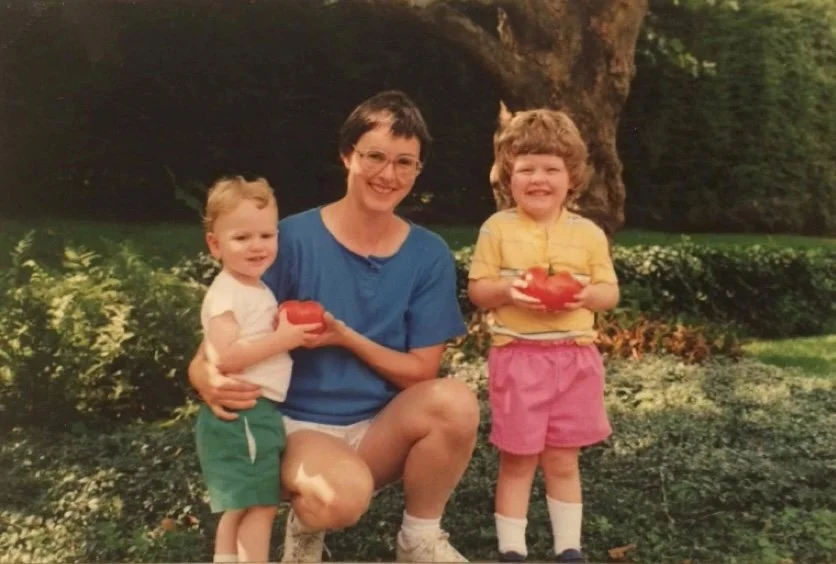 A woman with short dark hair and glasses sitting outdoors with two children, all holding tomatoes, surrounded by green bushes and trees.