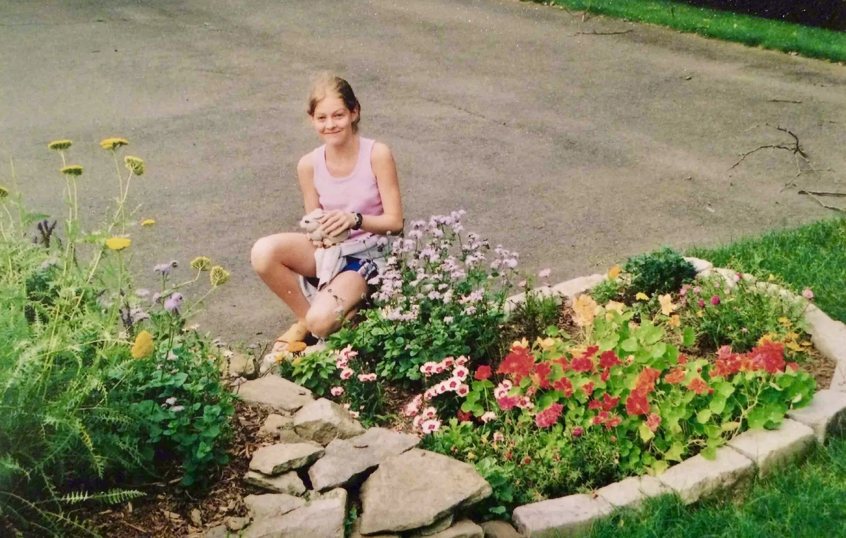 A young girl kneeling beside a flower bed, holding a small stone statue of a rabbit, with a driveway and a lawn in the background.