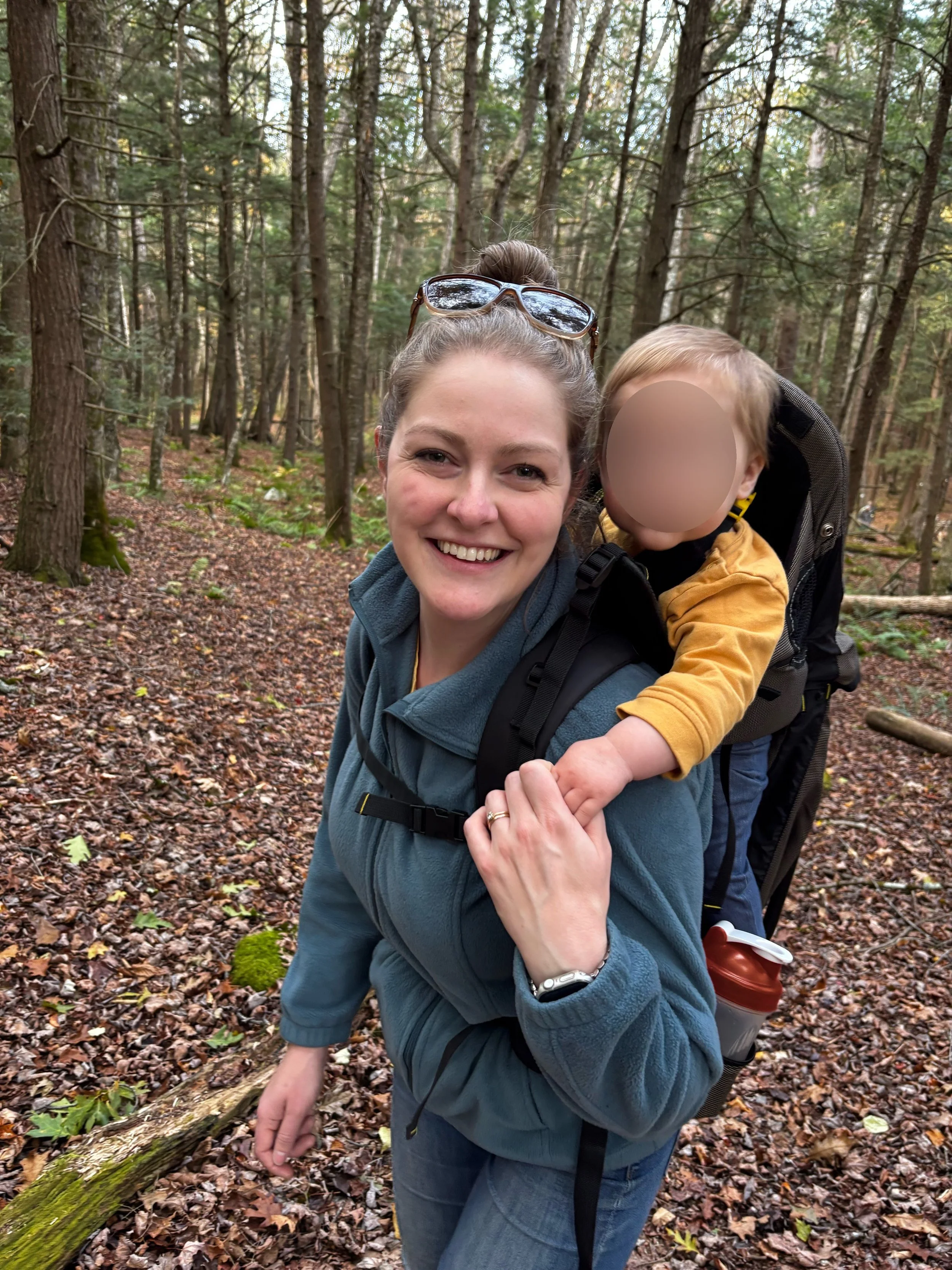 A woman is hiking in the woods wearing a backpack that carries a little boy