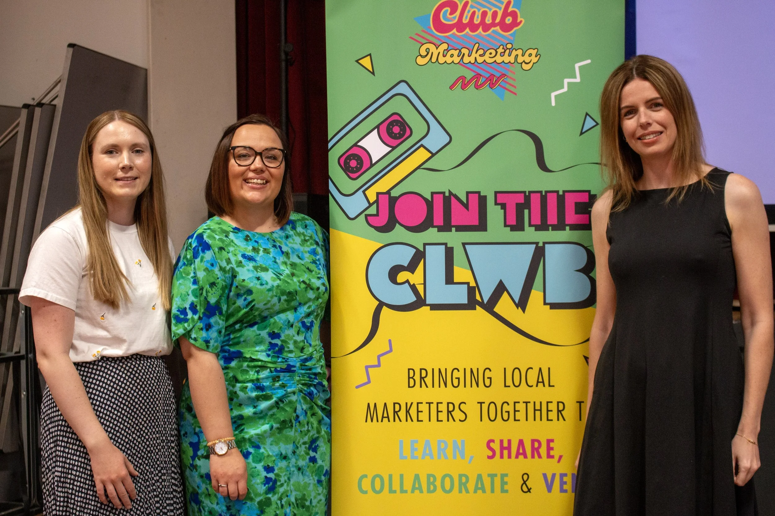 Three women standing in front of a colorful banner at a marketing event. The banner reads "Join the Club" with a cassette tape graphic and the text "Bringing local marketers together to learn, share, collaborate & vet." The women are smiling and dressed in casual to semi-formal attire.