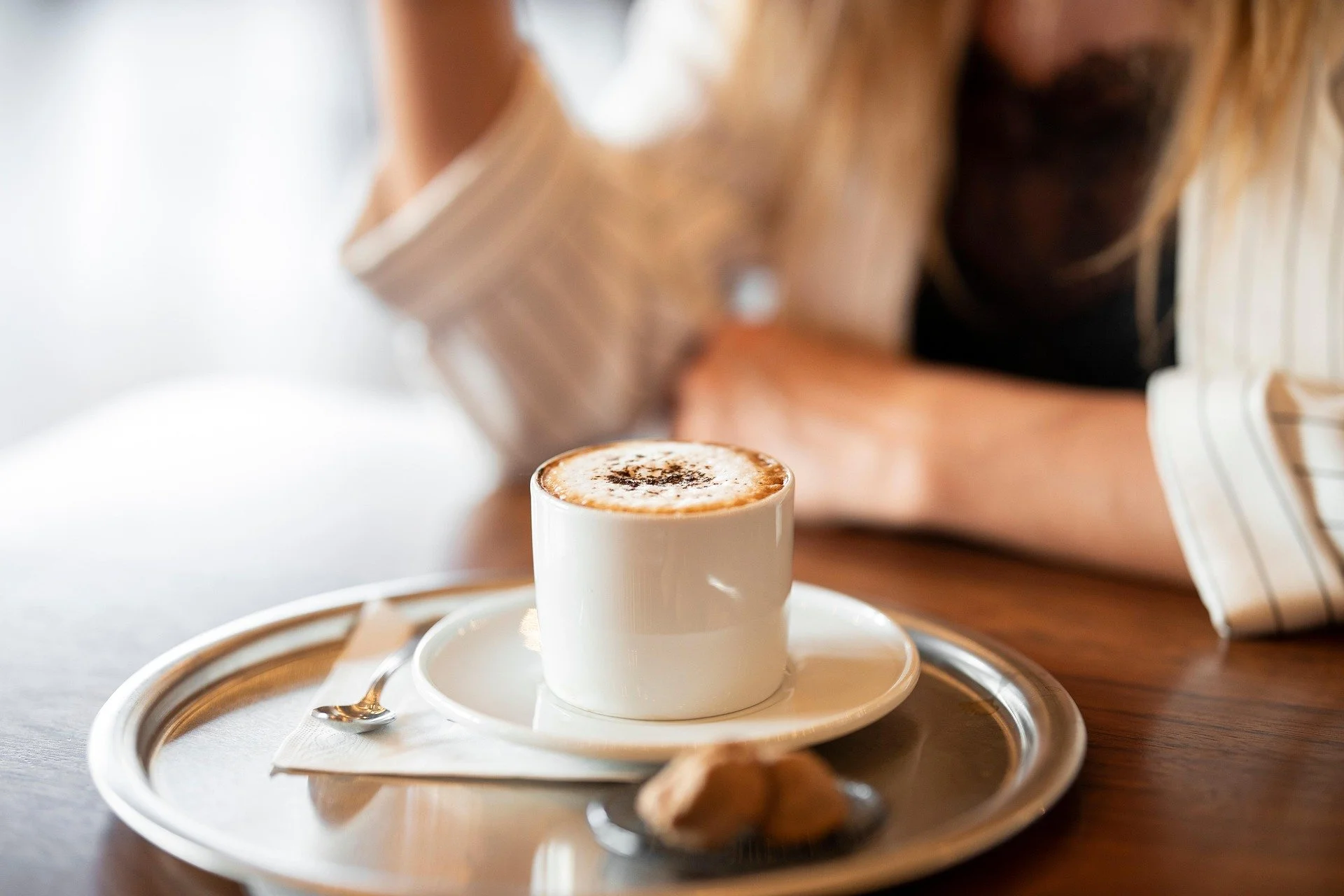 Cup of coffee on a white saucer with a spoon and biscuit on a silver tray, with a person sitting at a wooden table in the background.