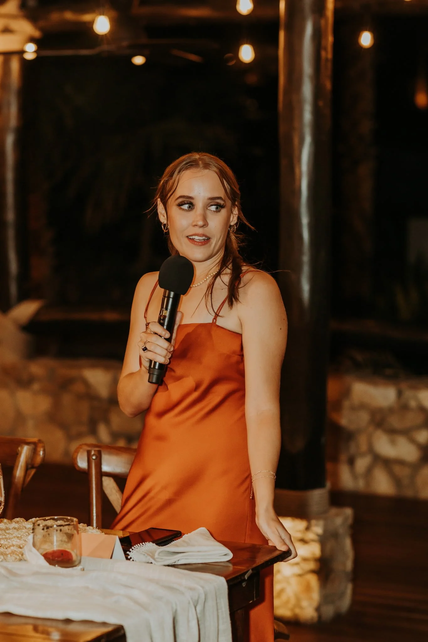 A woman in an orange satin dress holding a microphone speaks at a formal event in a rustic venue with hanging Edison bulbs and stone walls.