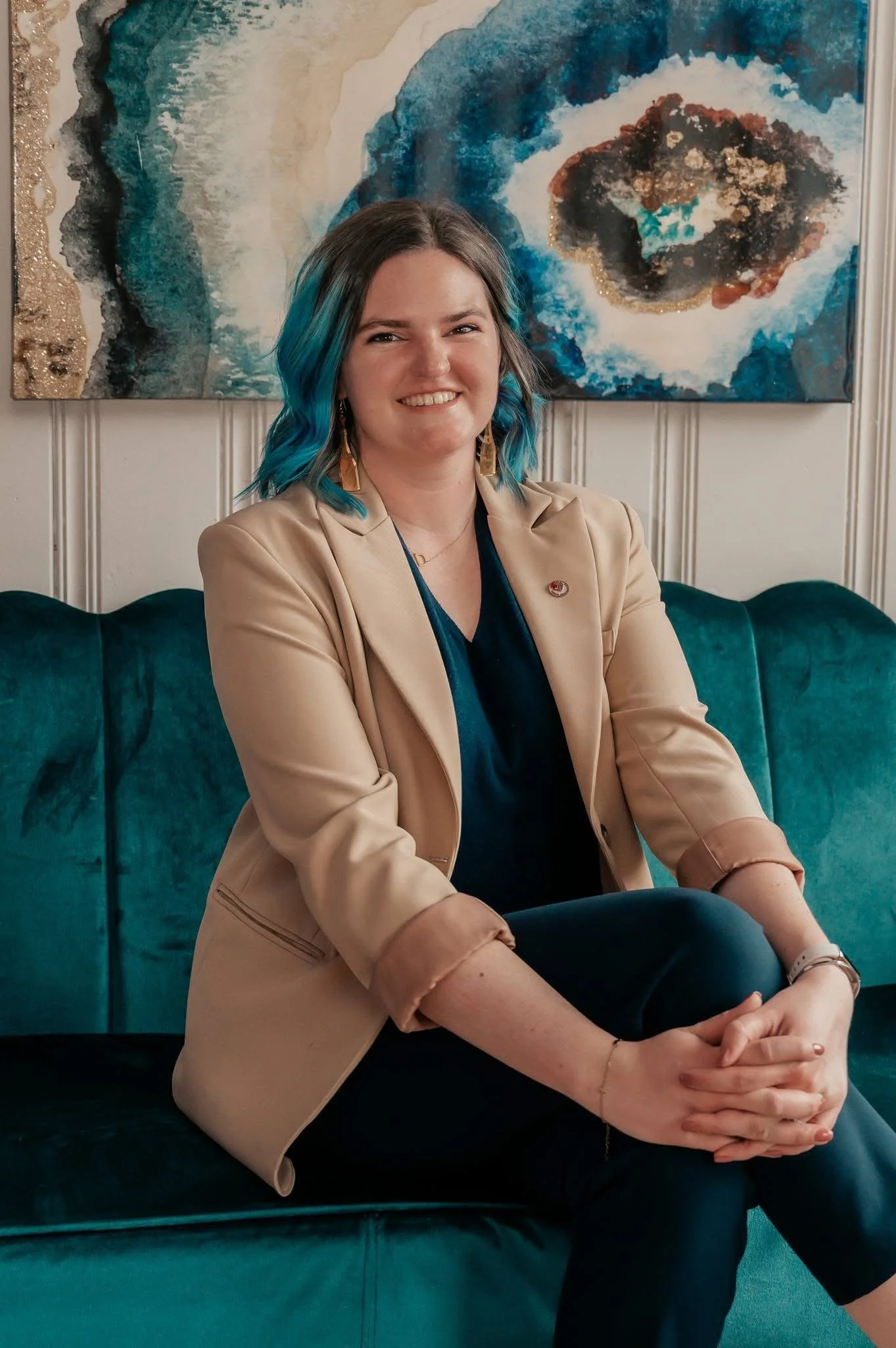A woman with shoulder-length hair featuring blue tips, wearing a beige blazer, navy top, and black pants, sitting on a dark green velvet couch with her hands clasped and smiling at the camera. Behind her is an abstract painting with blues, white, tan, and black colors.