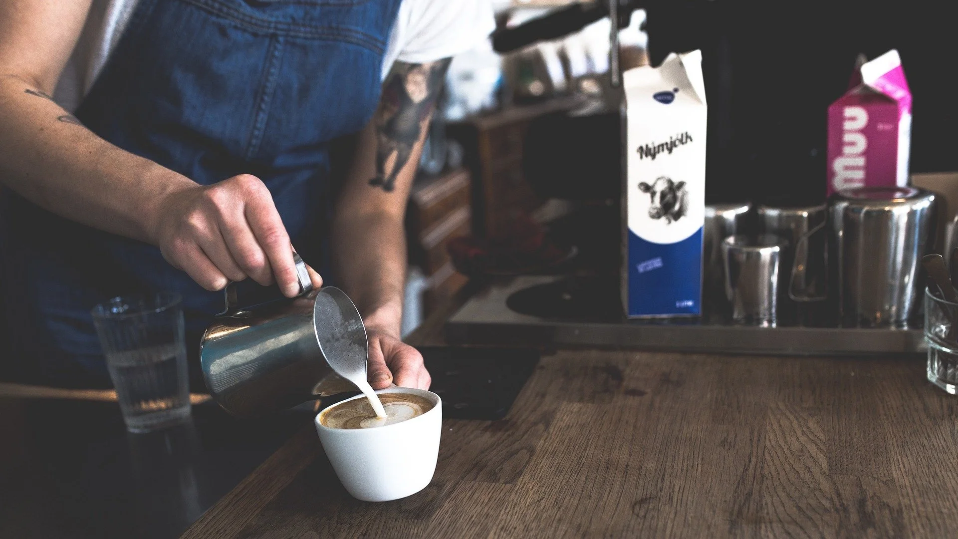 Person pouring steamed milk into a cup of coffee behind a wooden counter in a cafe.