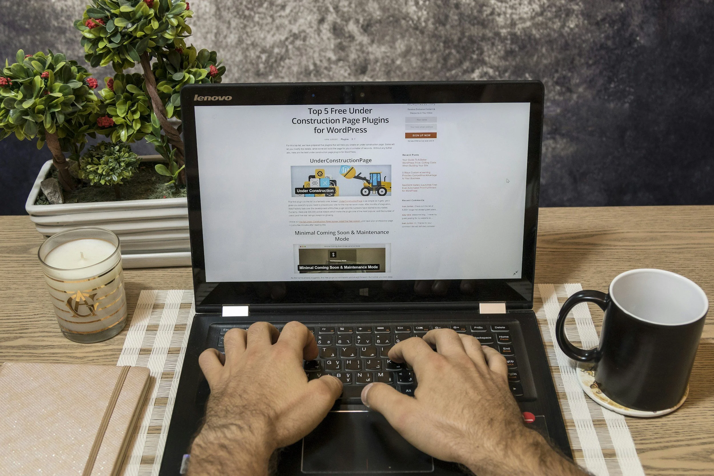 Person typing on a black Lenovo laptop at a wooden desk, with a white and gold candle on the left, a black and white mug on a coaster on the right, a pink notebook at the bottom left, and a potted plant with small green and red leaves in the background.