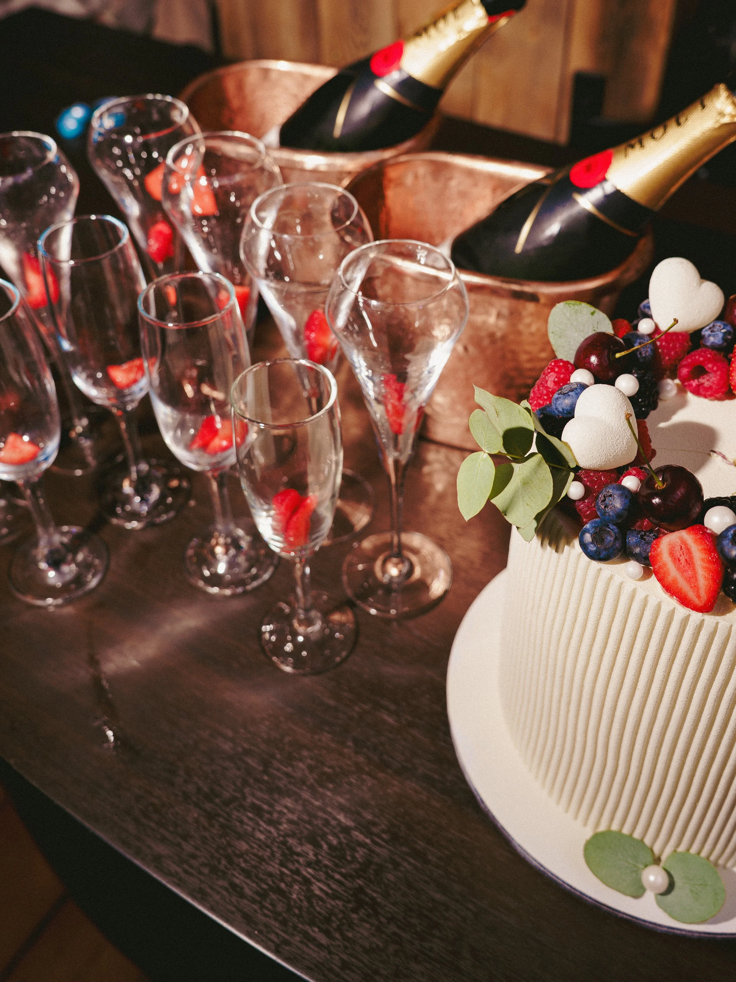 A celebration table with a white cake decorated with mixed berries and white hearts, champagne bottles chilling in ice buckets, and multiple empty crystal glasses with strawberries inside.