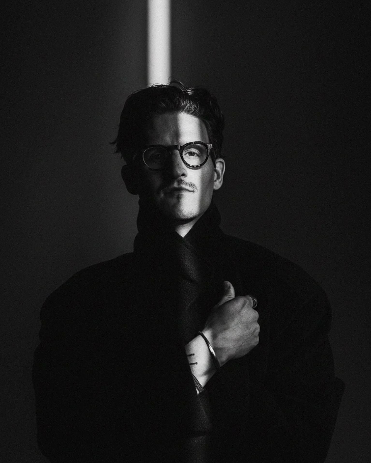 Black and white portrait of a man wearing glasses, with one hand over his chest, against a dark background with a vertical light streak.