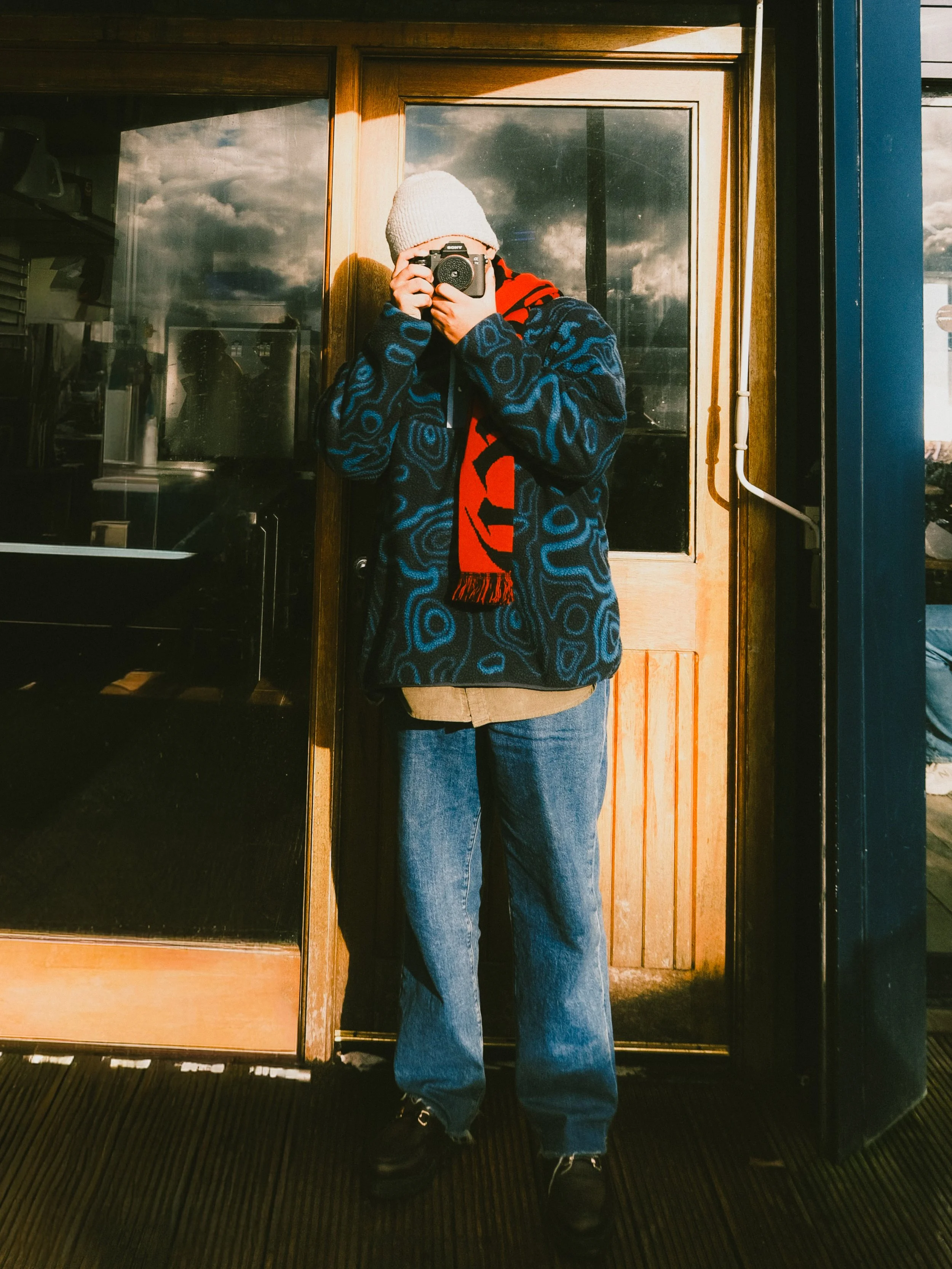 Person taking a selfie with a camera in front of a mirror, wearing a white knit hat, a navy patterned jacket, a beige shirt, blue jeans, and black shoes, standing outside near a wooden door and glass window.
