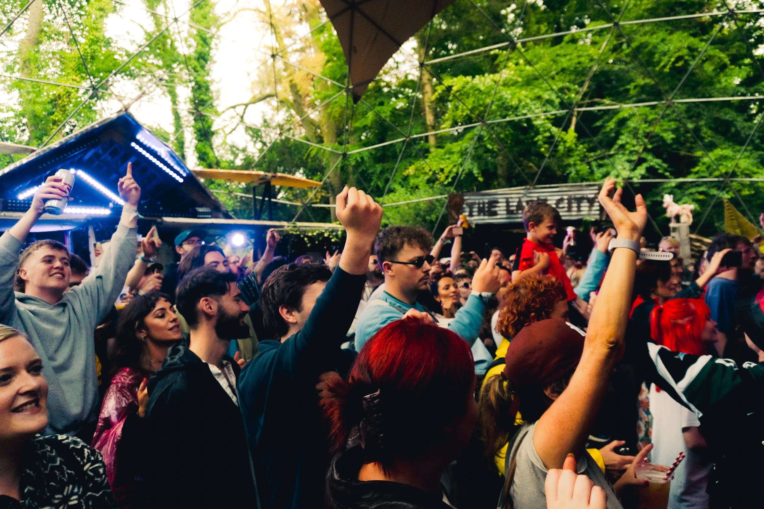 Crowd of people at an outdoor concert or festival, some with raised hands, surrounded by trees and canopy, with a stage and sign in the background.