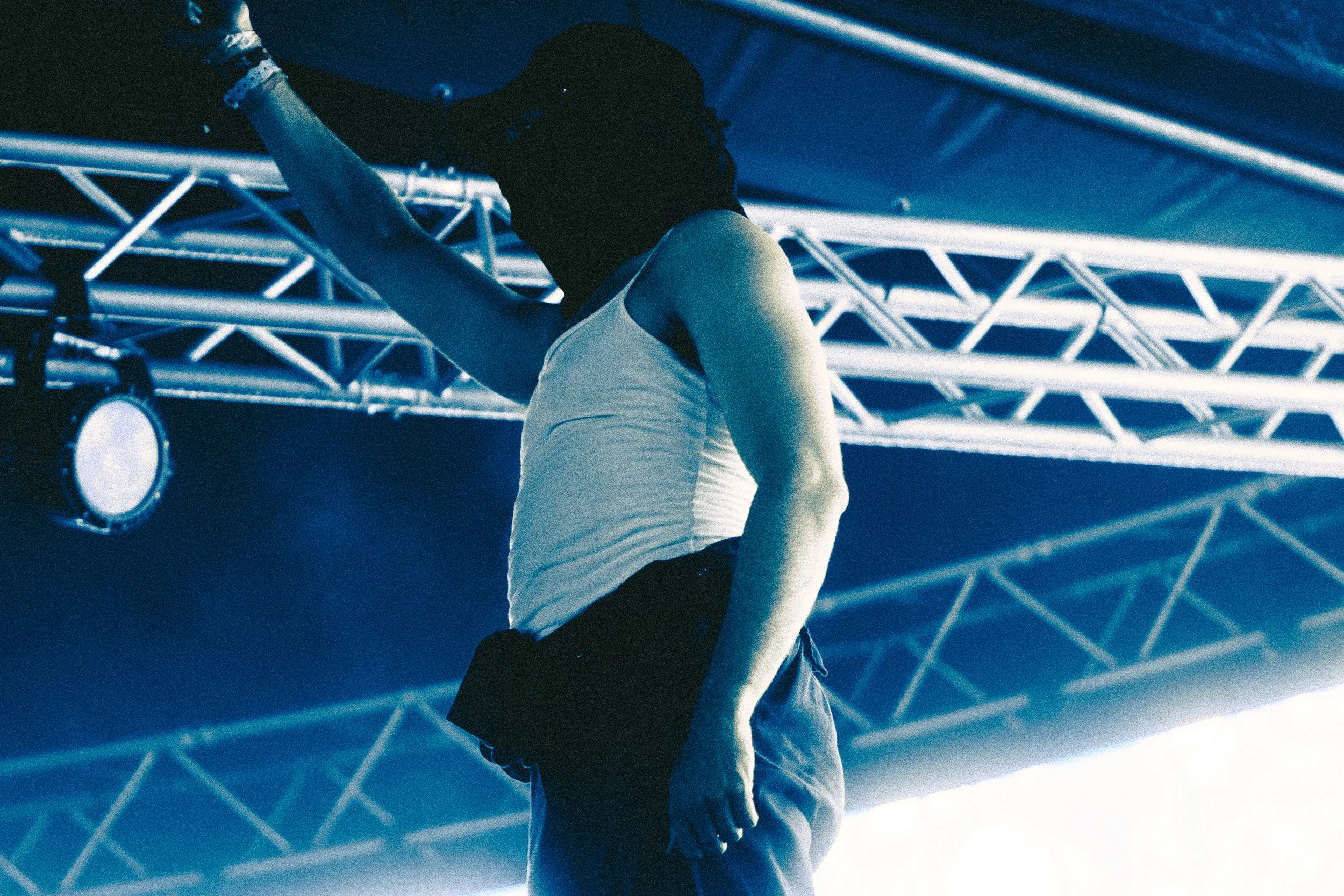 A male performer on stage, dressed in a white tank top and dark pants, with one arm raised and the other arm down, under stage lights and metal truss structure.