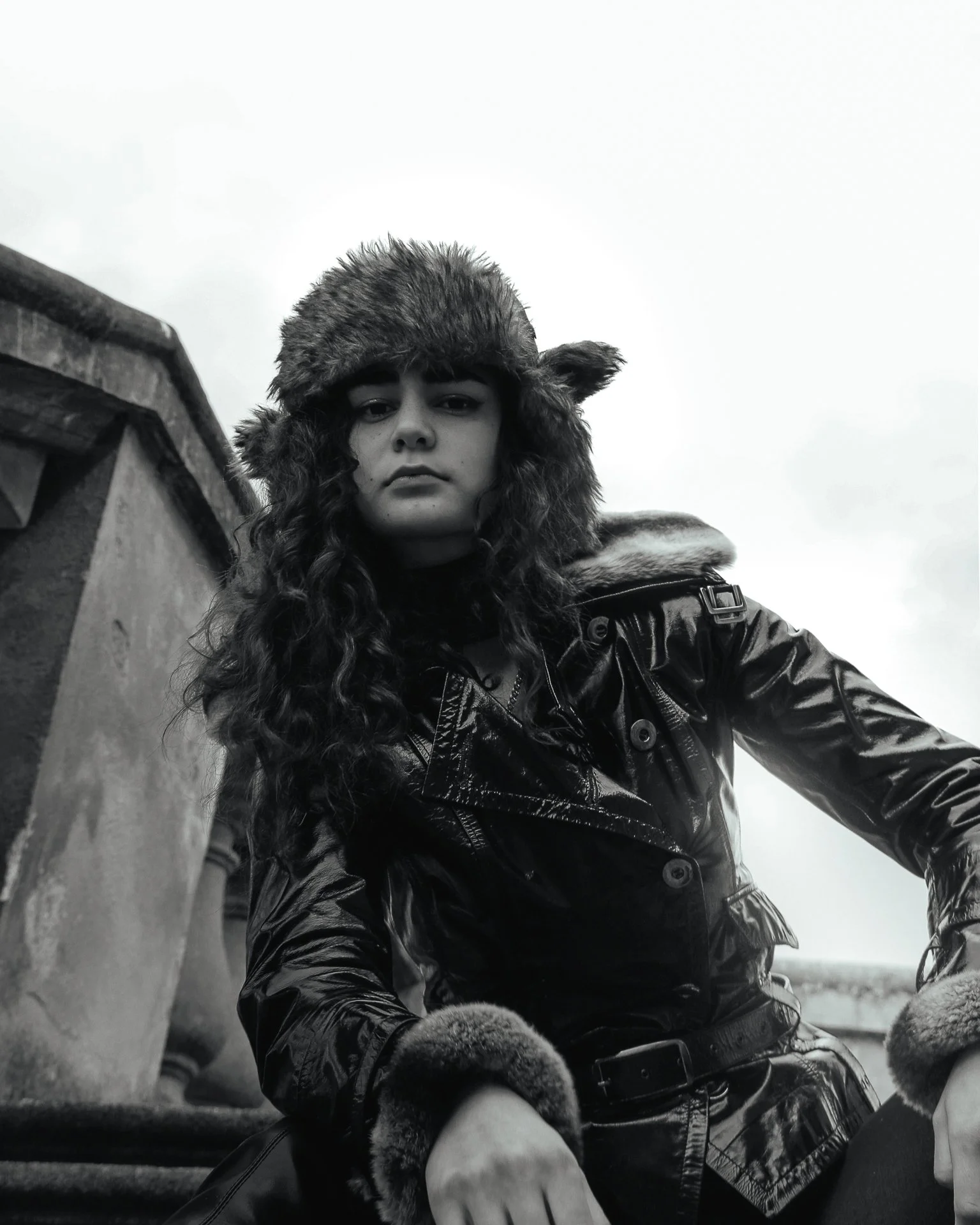 A young woman with wavy hair wearing a fur hat with ear flaps, a shiny black leather jacket with a fur collar, and fur cuffs on her sleeves, posing outdoors against a cloudy sky and rooftop.