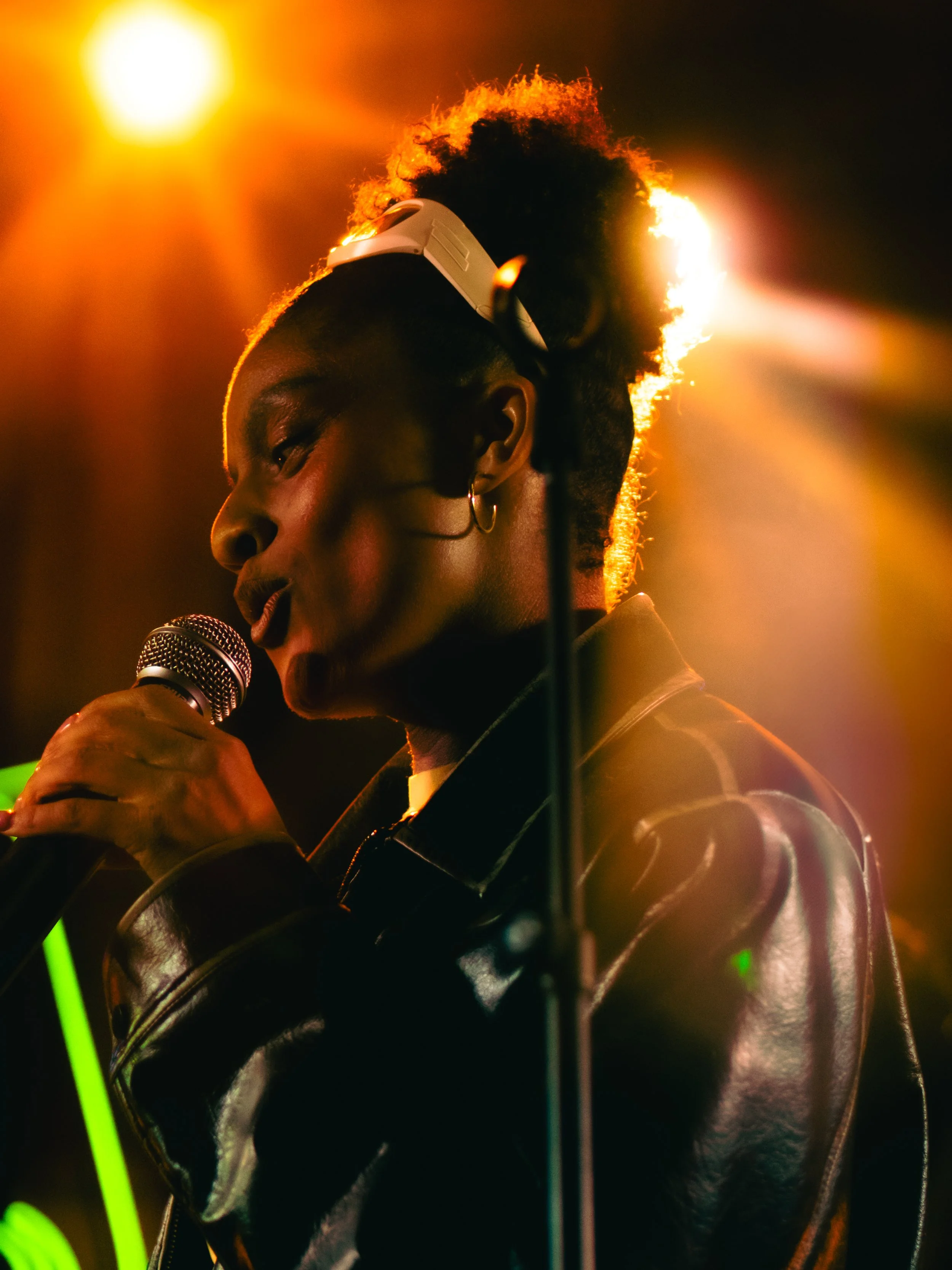 A woman with curly hair tied up, wearing headphones, singing into a microphone on stage with warm lighting.