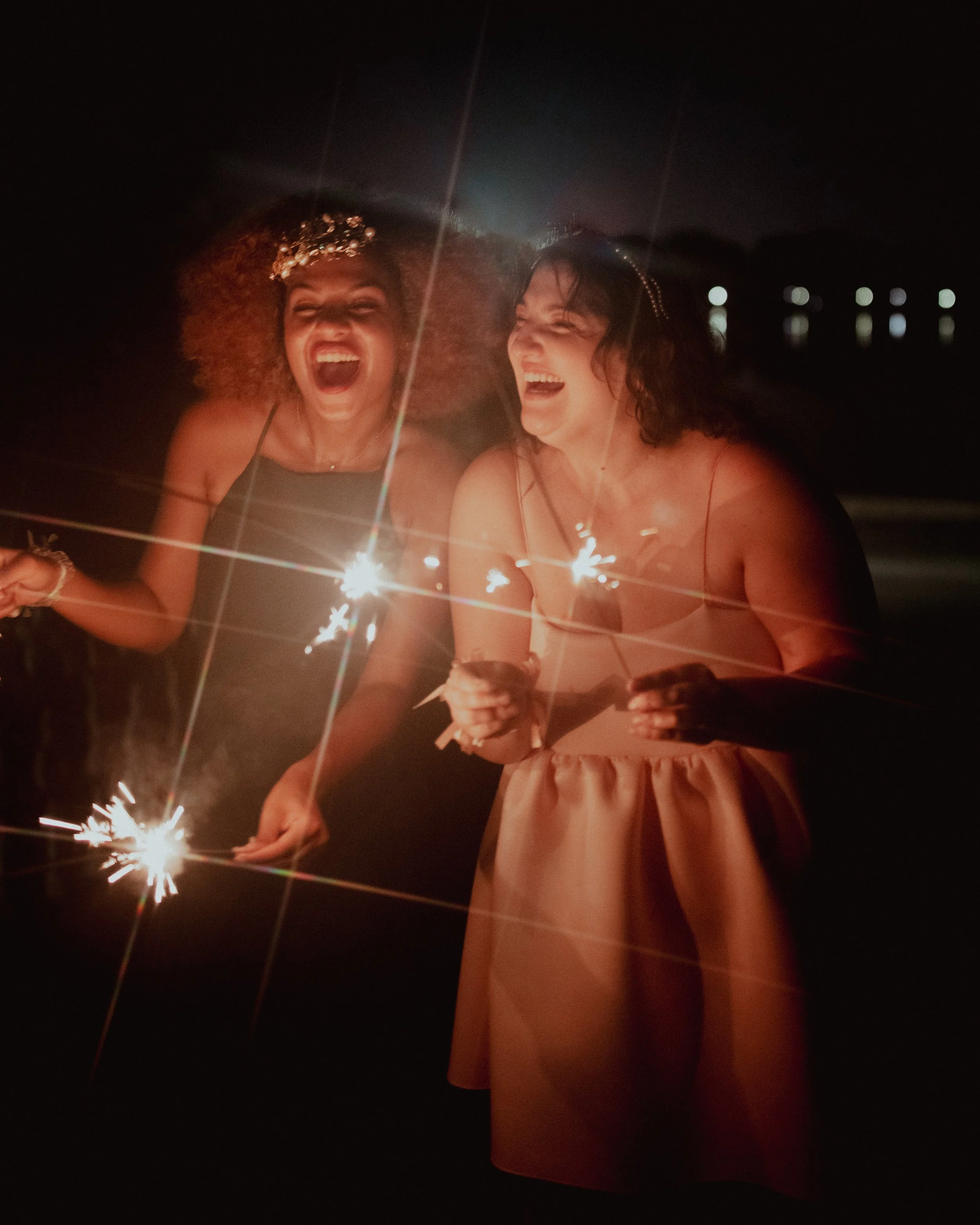 Two women in dresses holding sparklers and laughing on a beach at night.