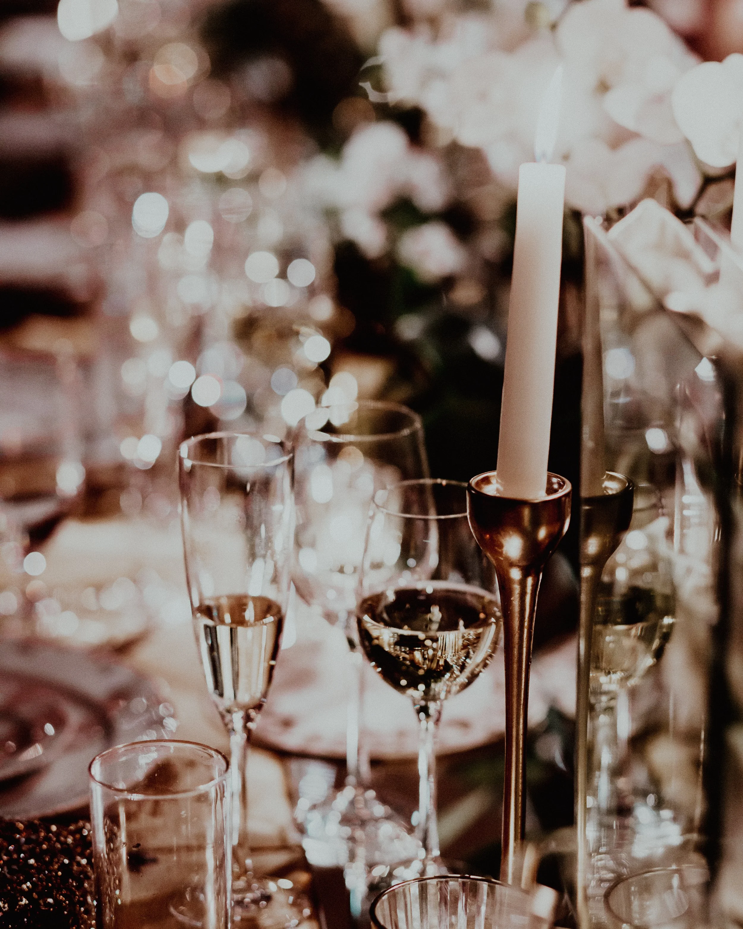 Set dining table with champagne flutes, wine glasses, and several candle holders, including a tall white candle in a gold holder, with soft lighting and floral decorations in the background.