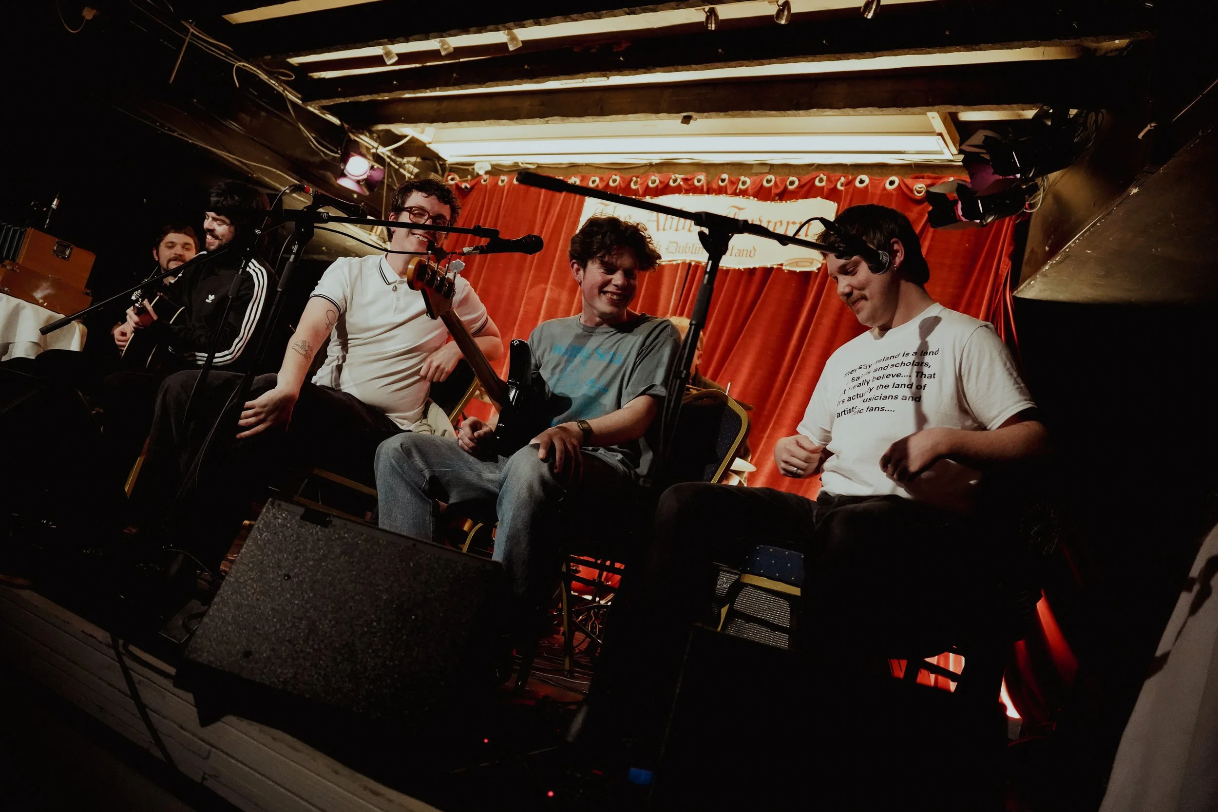 Group of young musicians performing on a stage with red curtains, playing guitars and singing into microphones, warmly lit, at an indoor venue.