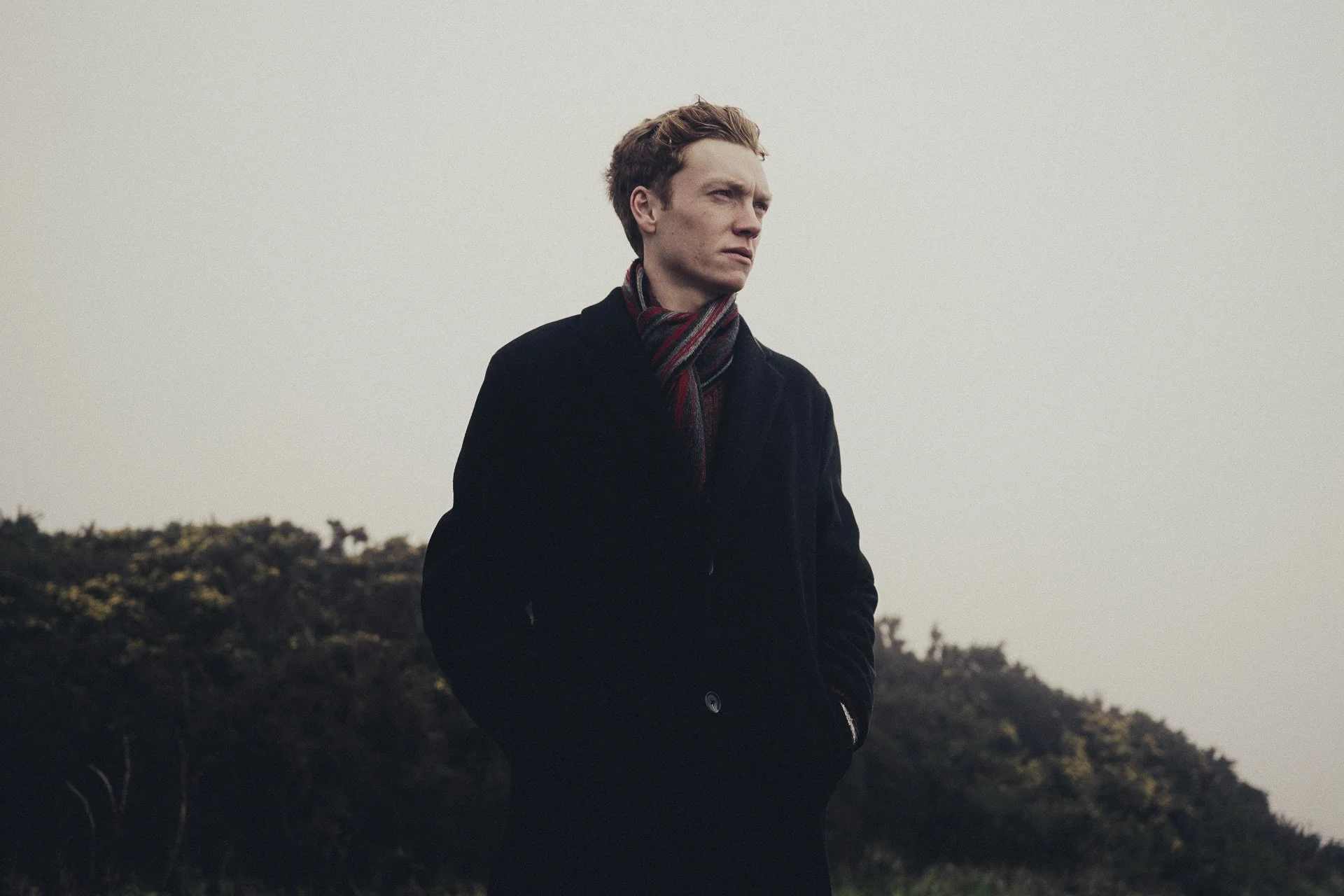 A young man standing outdoors on a cloudy day, looking to the side with a thoughtful expression. He is wearing a black coat and a striped scarf, with his hands in his pockets, standing in front of a hilly landscape.