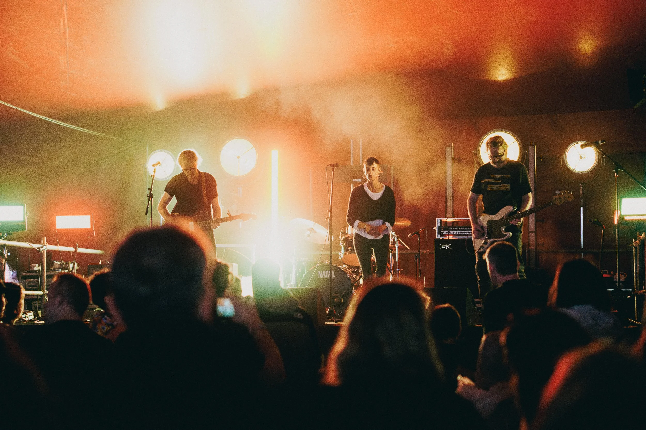 A band of three musicians performs on stage at a concert. The stage is illuminated with bright orange and yellow lights, with the audience visible in the foreground.