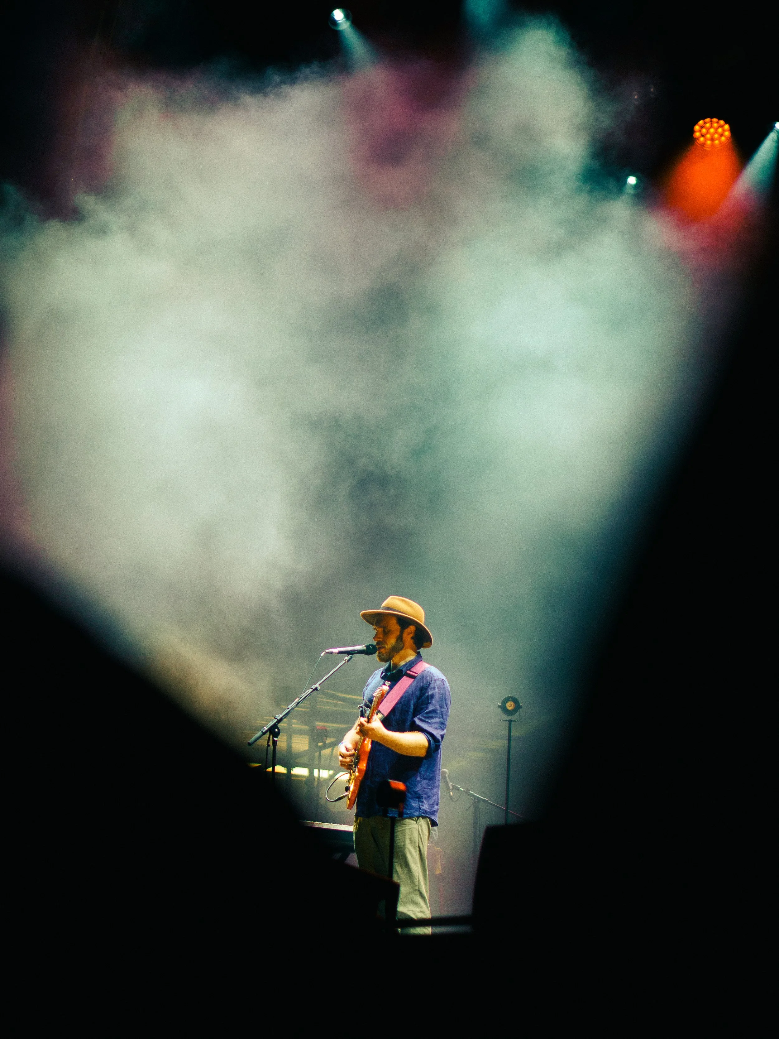 Male musician wearing a hat, playing guitar and singing into a microphone on stage, with colorful stage lights and fog.