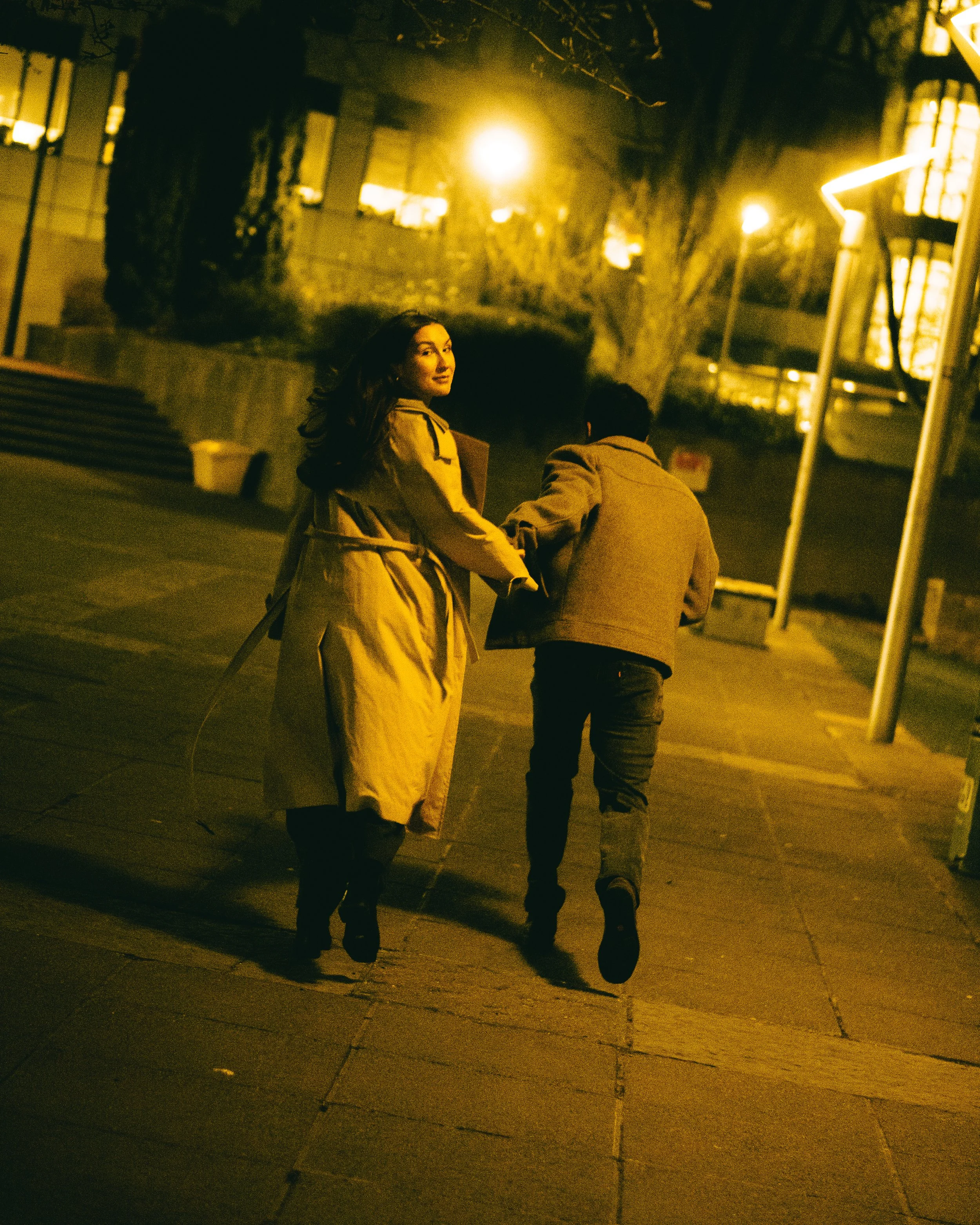 A woman and a man are walking together at night on a city sidewalk, holding hands or linked. The woman is looking back and smiling, wearing a yellow coat, while the man is turned away, dressed in a brown jacket.