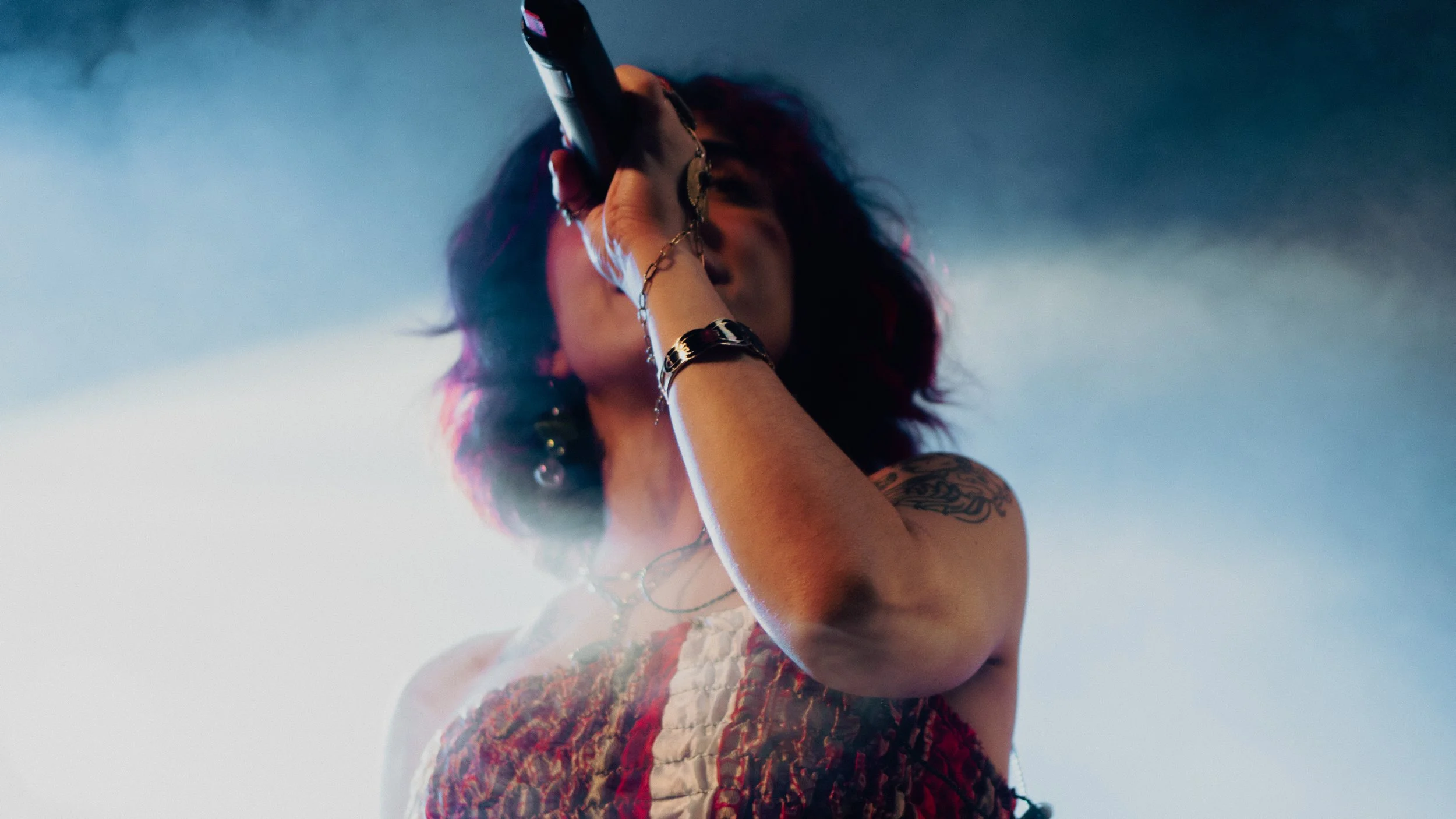 A female singer with dark curly hair singing into a microphone on stage, wearing jewelry and a patterned dress.