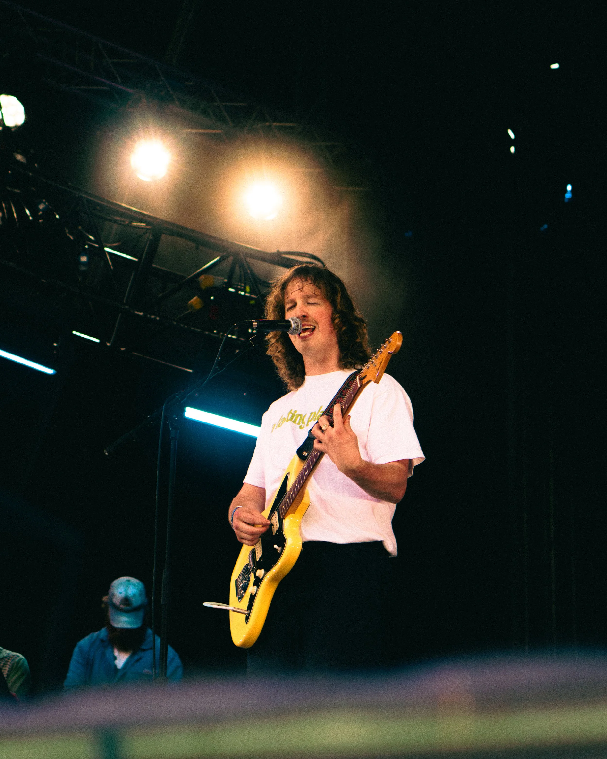 A musician singing and playing yellow electric guitar on stage under bright stage lights, wearing a white t-shirt.