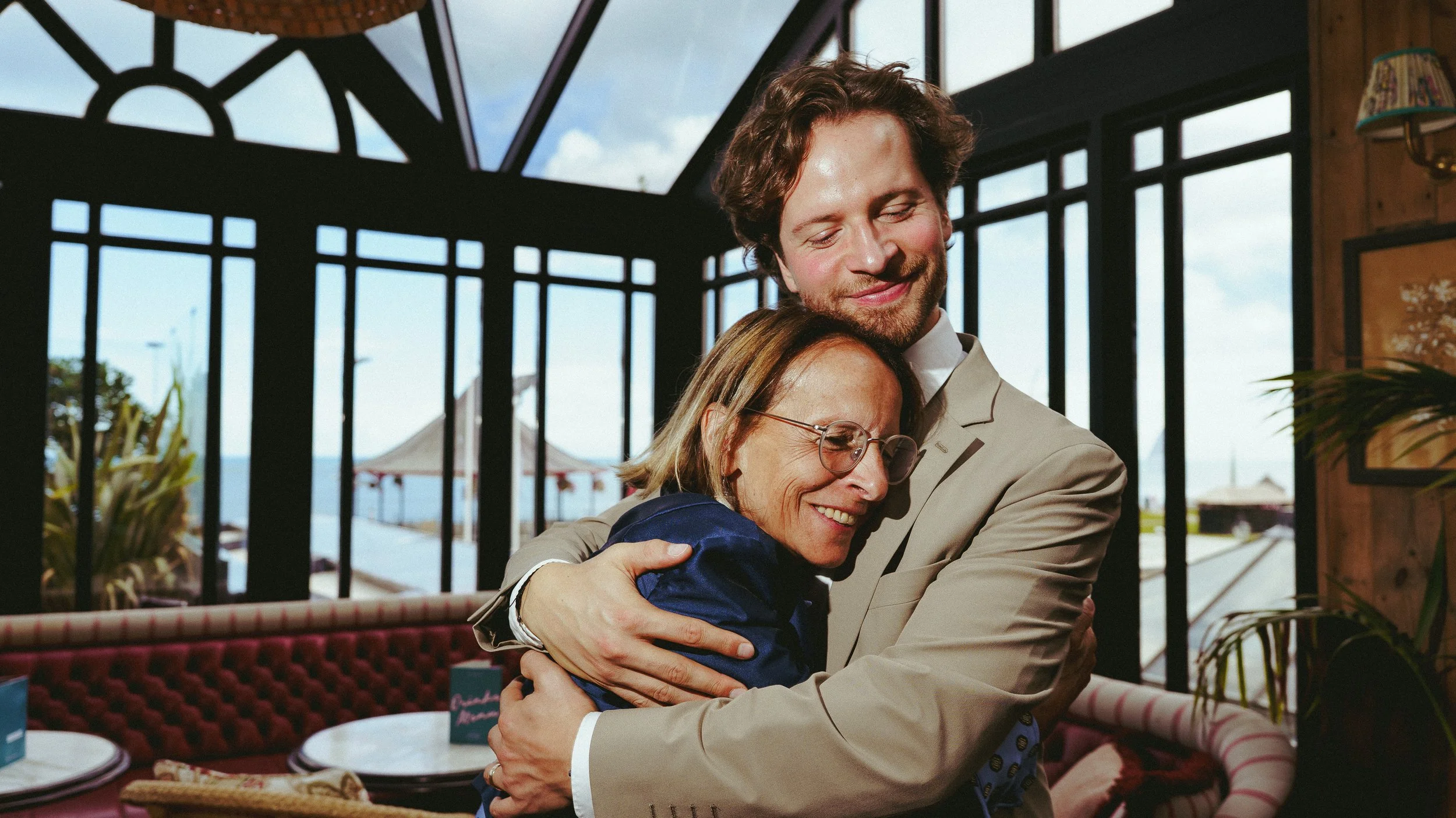 A woman with glasses and a man are hugging and smiling inside a restaurant or cafe with large windows and a view of the ocean outside.