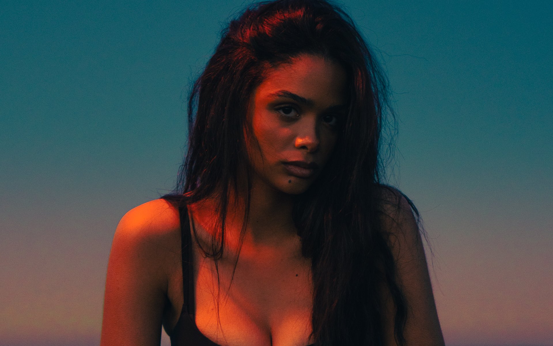 A woman with long, dark, wet hair looking directly at the camera against a gradient sky background.