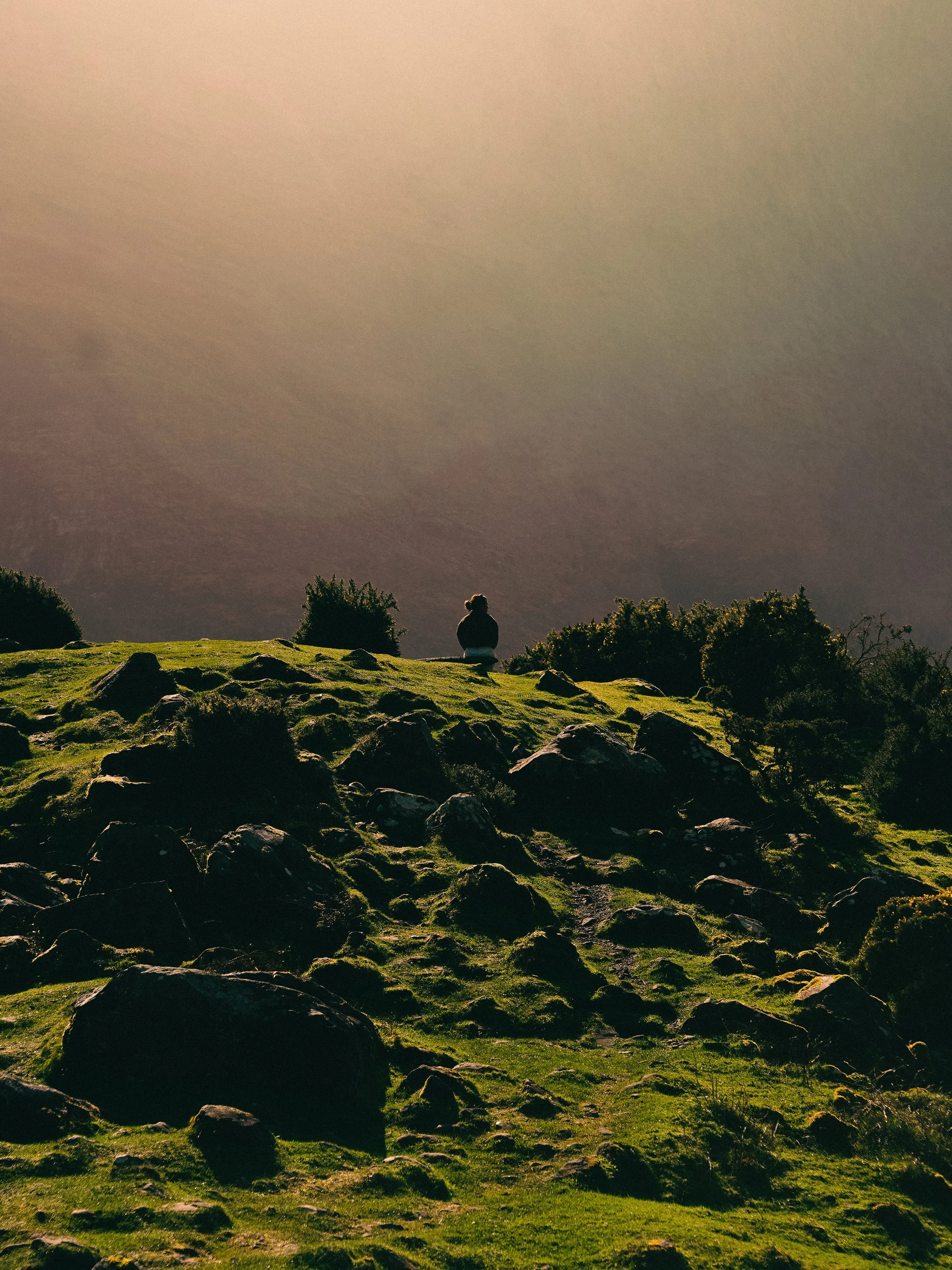 A person sitting on a grassy hilltop with rocks, facing away, under a cloudy sky during sunset.
