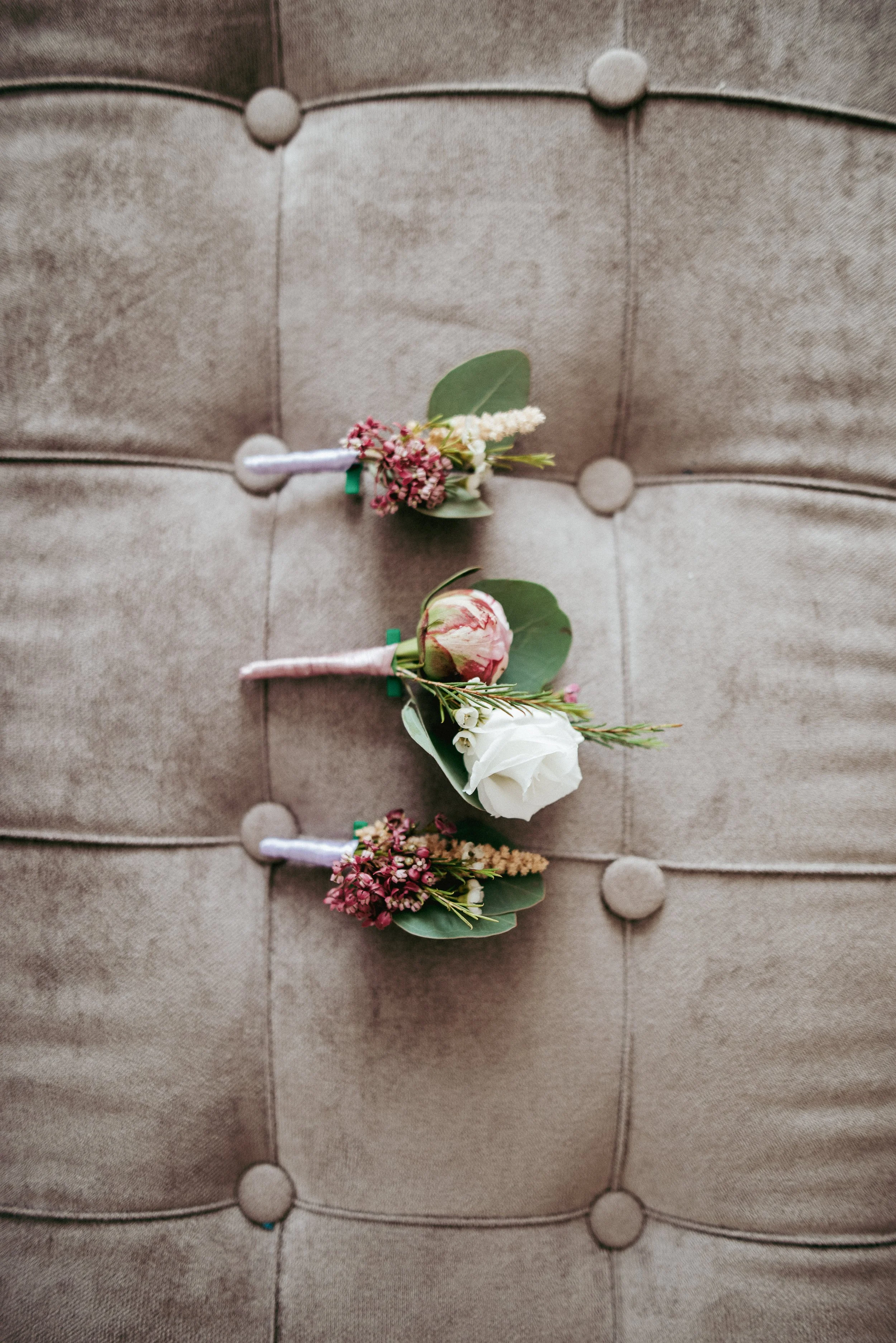 Three floral boutonnieres with pink, white, and green flowers on a beige tufted fabric surface.