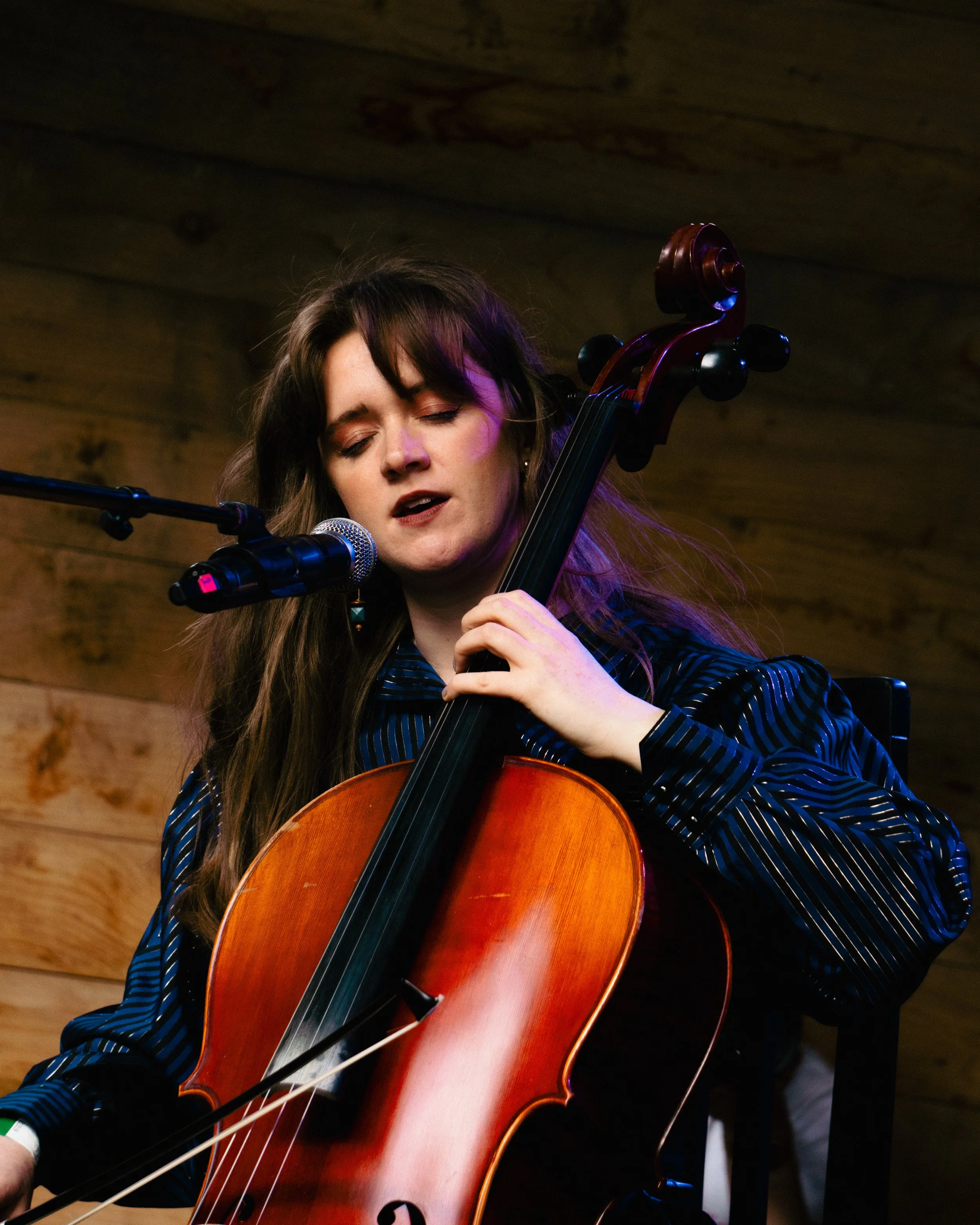 A woman with long brown hair singing and playing the cello at a microphone, with a wooden wall background.