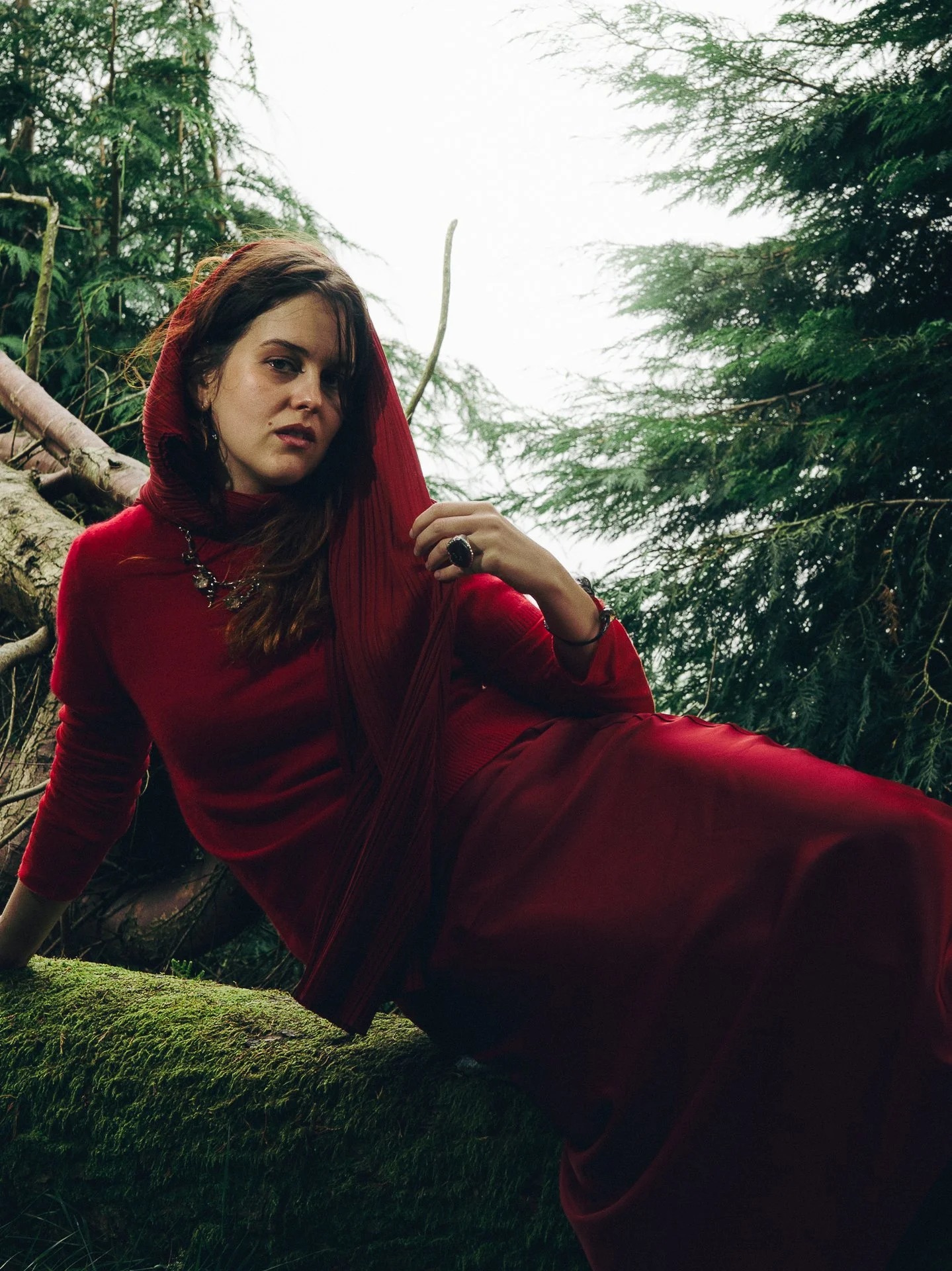 A woman in a red dress and hood sitting on a moss-covered fallen tree in a forest with green trees and cloudy sky in the background.