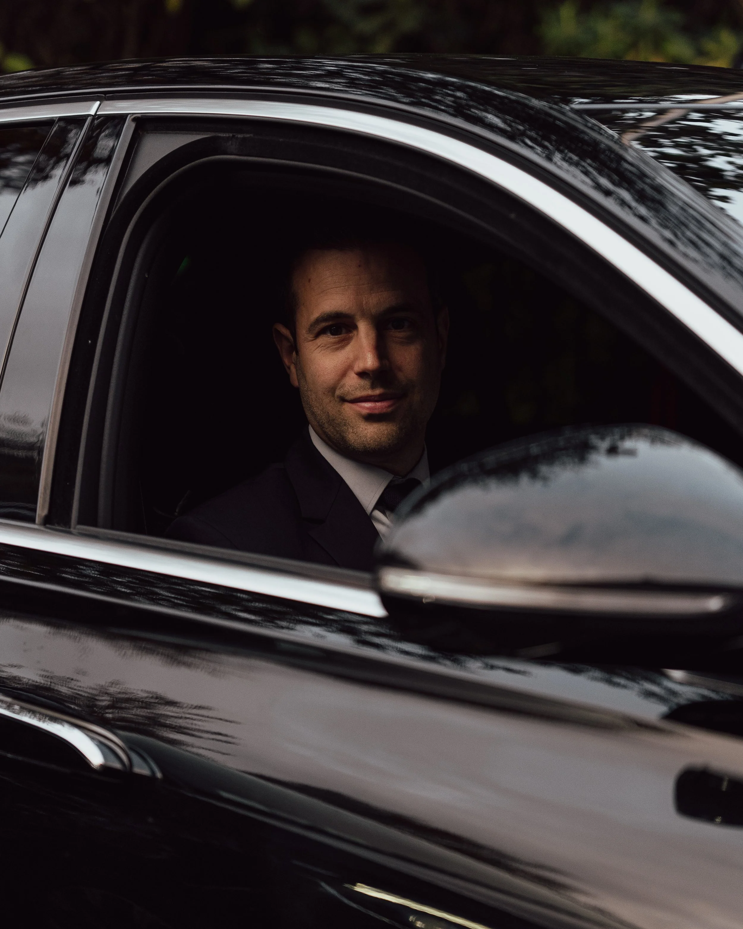 A man in a suit sitting in the driver seat of a black car, smiling at the camera.