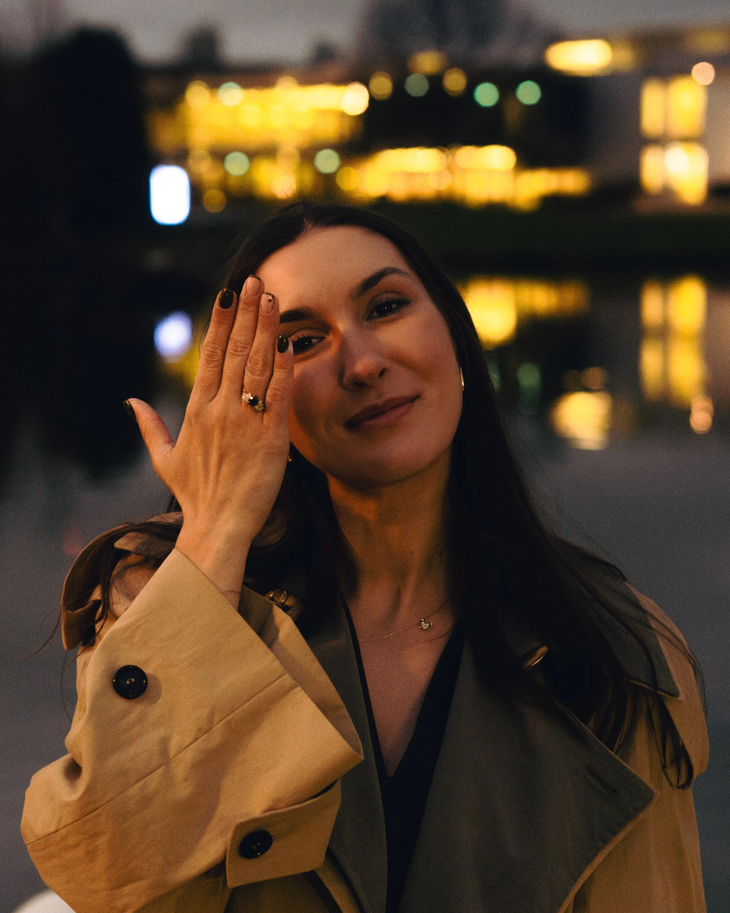 A woman in a tan trench coat smiling and touching her face with her hand, standing outdoors at night with city lights reflecting on water behind her.