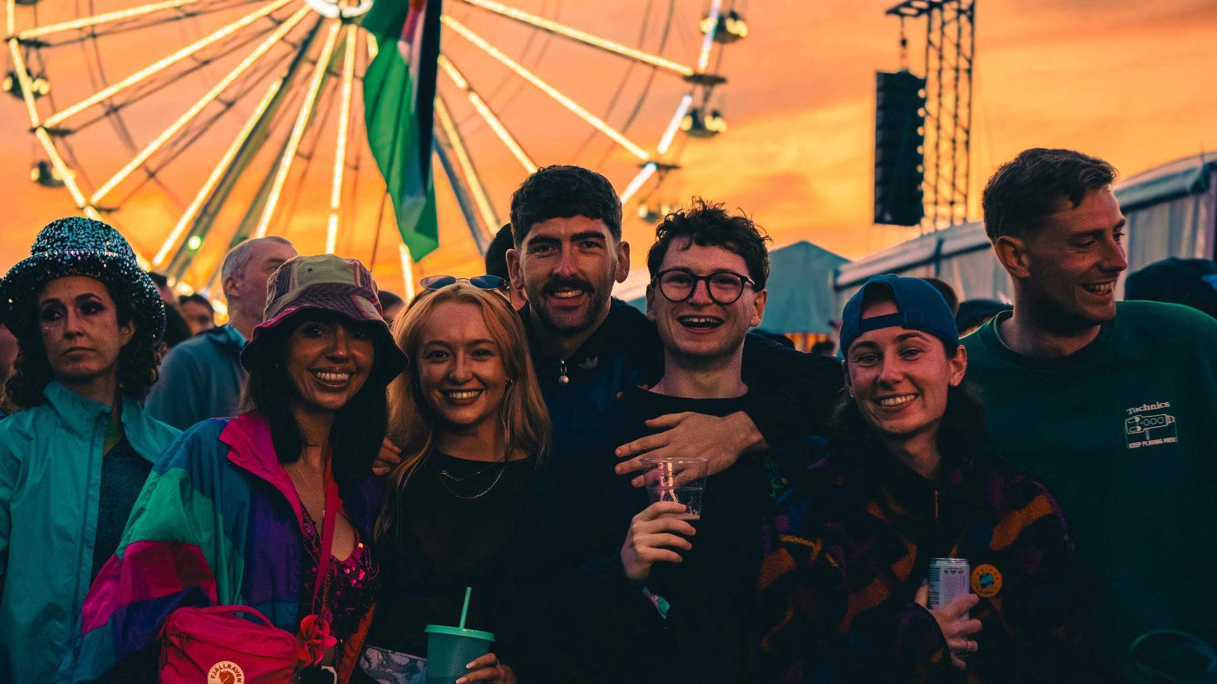 A group of smiling people at a carnival with a ferris wheel and sunset in the background.